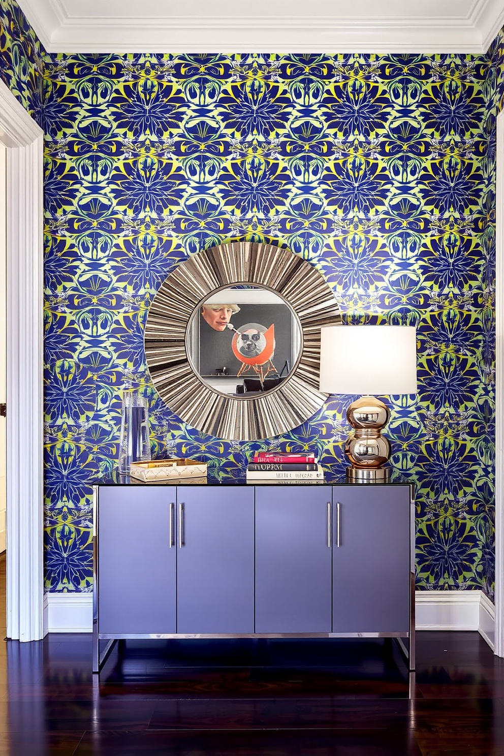 A bold wallpaper featuring a vibrant geometric pattern covers the statement wall in the apartment entryway. The entryway is enhanced by a sleek console table adorned with a decorative mirror and a stylish lamp, creating an inviting atmosphere.