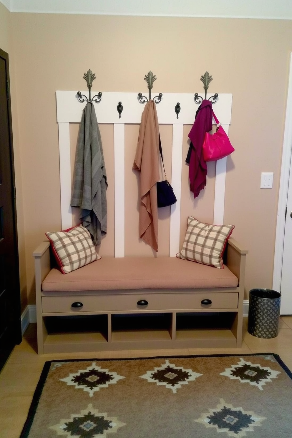 A multi-purpose entryway bench with soft cushions sits against the wall. The bench features built-in storage underneath and is flanked by decorative hooks for hanging coats and bags. The walls are painted in a warm neutral tone, creating an inviting atmosphere. A stylish area rug lies beneath the bench, adding texture and warmth to the space.