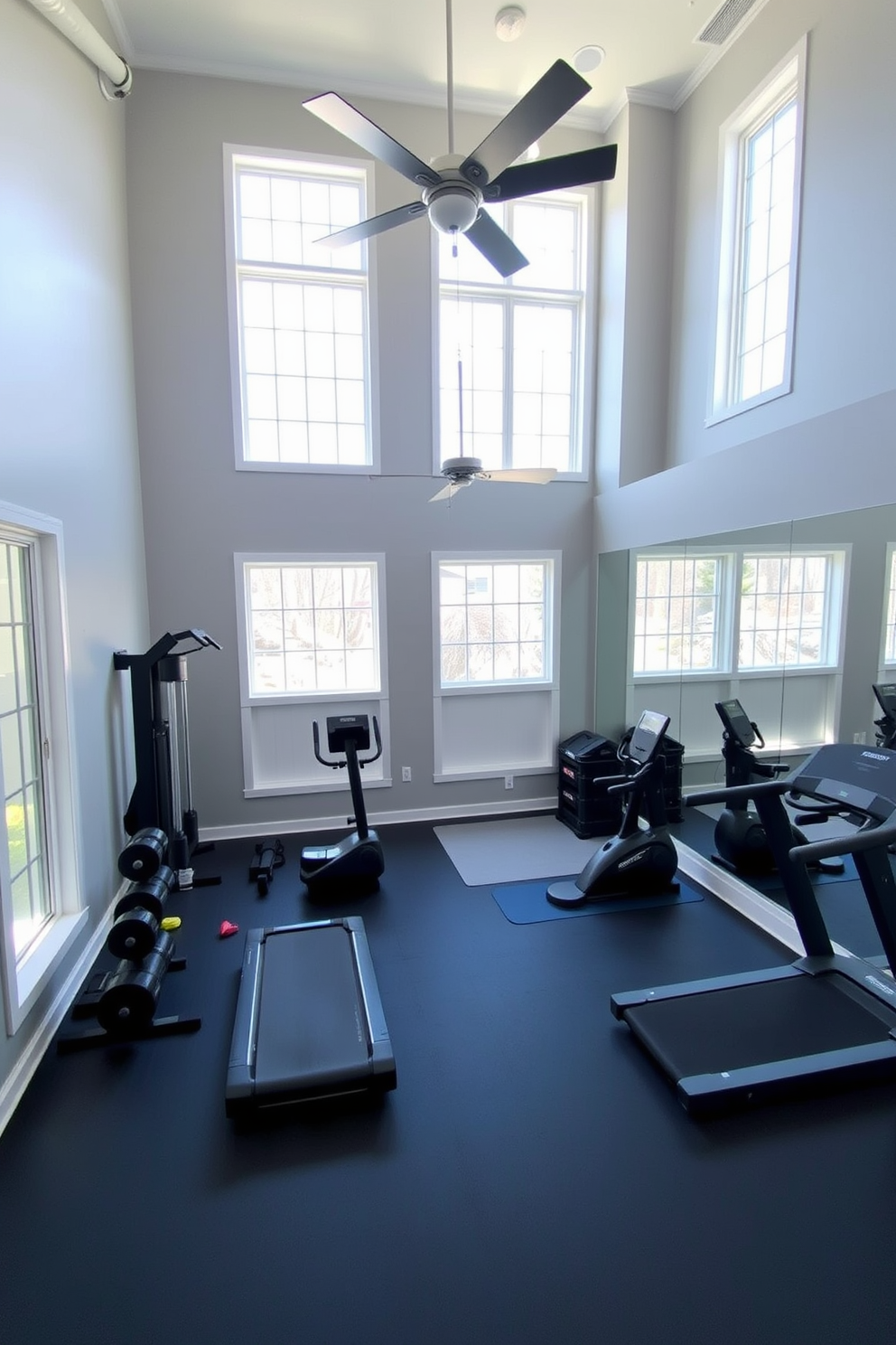 A stylish basement gym featuring high ceilings and large windows that let in natural light. The space includes a variety of workout equipment such as free weights, a treadmill, and a yoga area with mats. The walls are painted in a calming light gray, and the flooring is a durable rubber material for safety. A ceiling fan provides ventilation and comfort while a large mirror reflects the space, making it feel more open and inviting.