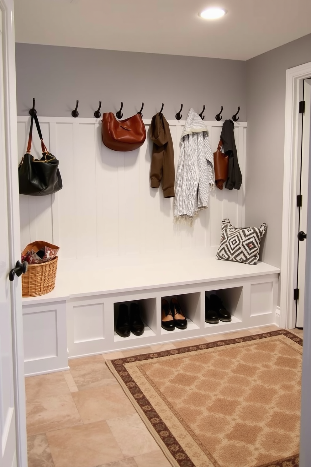 A functional basement mudroom featuring a spacious built-in bench with storage cubbies underneath. The walls are painted in a soft gray tone, and the floor is covered with large ceramic tiles for durability. A cozy area rug in a geometric pattern adds warmth and style to the space. Hooks for coats and bags line the wall above the bench, while a decorative basket holds shoes neatly out of sight.