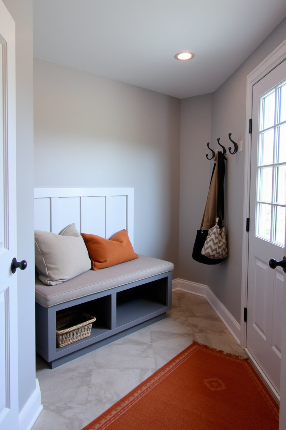 A cozy basement mudroom featuring a built-in bench with storage underneath. The walls are painted in a soft gray tone, and the floor is covered with a durable, patterned tile in neutral shades. To the right, a row of hooks is mounted for hanging coats and bags, while a large window allows natural light to brighten the space. A decorative rug in warm earth tones adds comfort and style underfoot.