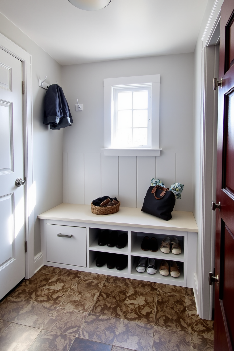 A cozy mudroom features a built-in bench with ample shoe storage underneath. The walls are painted a soft gray, and the floor is covered with durable, patterned tiles for easy cleaning. On one side of the bench, there are hooks for hanging coats and bags. Natural light streams in through a small window, creating a welcoming and functional space.