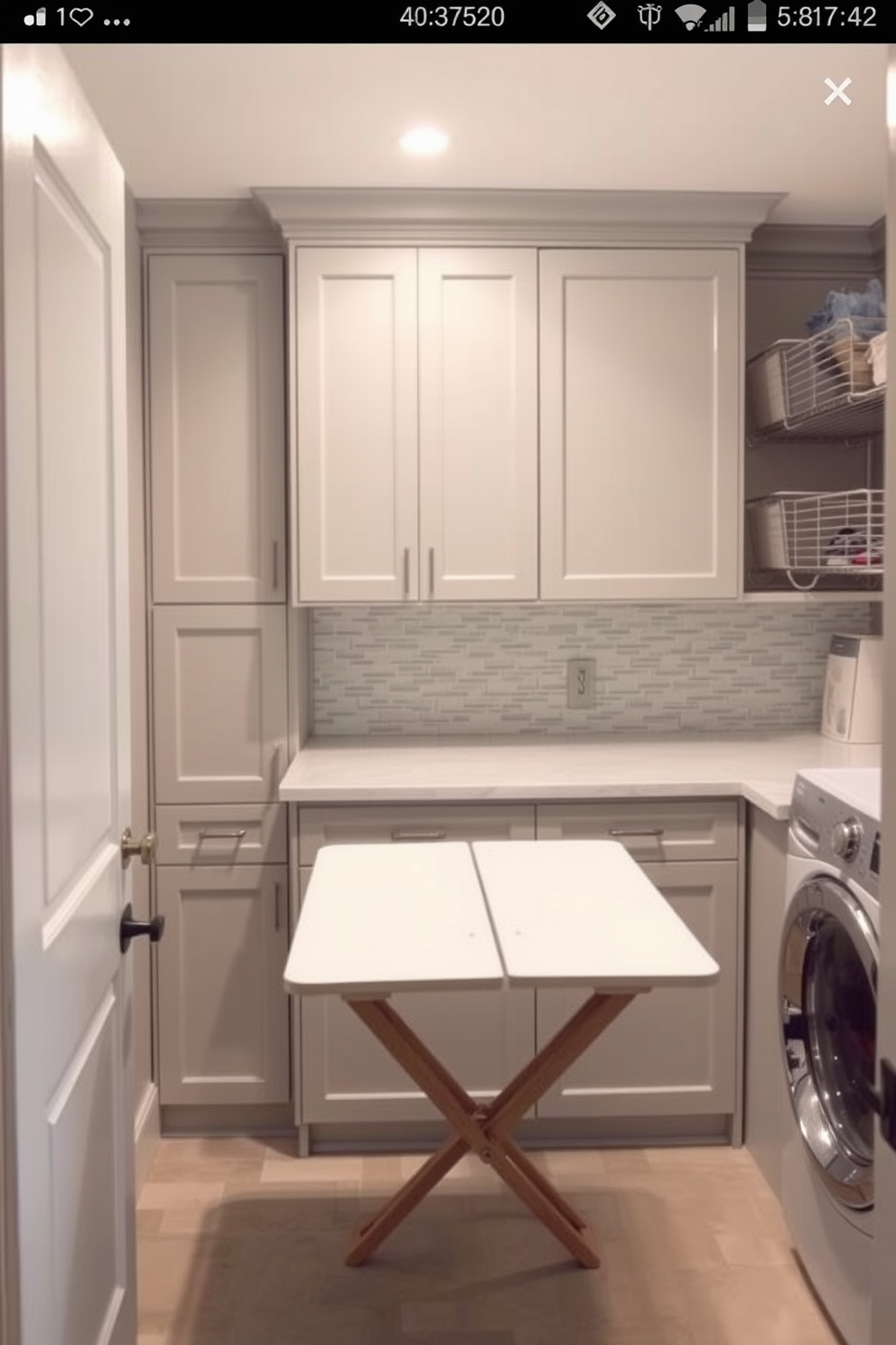 A functional laundry room featuring a folding table adjacent to a washer and dryer. The space is designed with light gray cabinetry and a stylish backsplash, creating a modern yet practical environment.