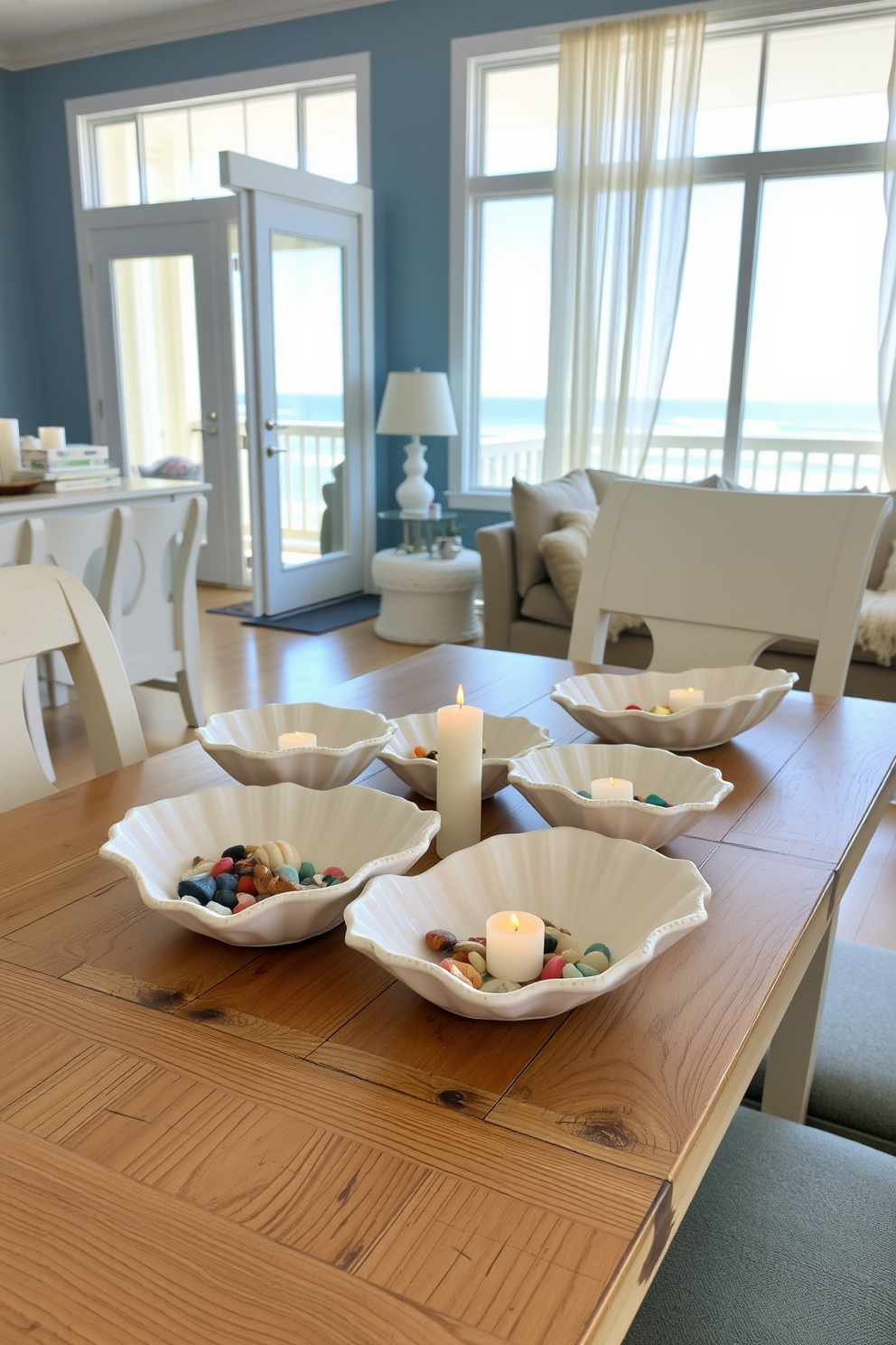 A stylish beach apartment setting featuring ceramic seashell dishes arranged on a wooden dining table. The dishes are filled with colorful beach stones and small candles, creating a relaxed coastal vibe. The walls are painted in a soft blue hue, complementing the whitewashed wooden furniture. Large windows allow natural light to flood the space, showcasing ocean views and airy sheer curtains.