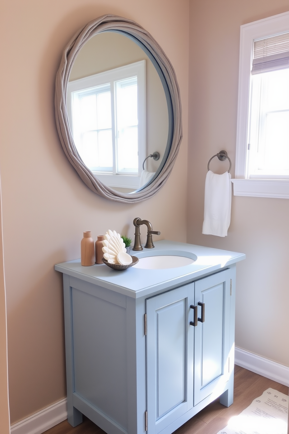 A simple vanity with a coastal finish features a light blue color palette and natural wood accents. The countertop is adorned with a seashell-inspired decor piece and a small potted plant for a fresh touch. The walls are painted in a soft sandy beige, creating a warm and inviting atmosphere. A large round mirror with a driftwood frame hangs above the vanity, reflecting the natural light from a nearby window.