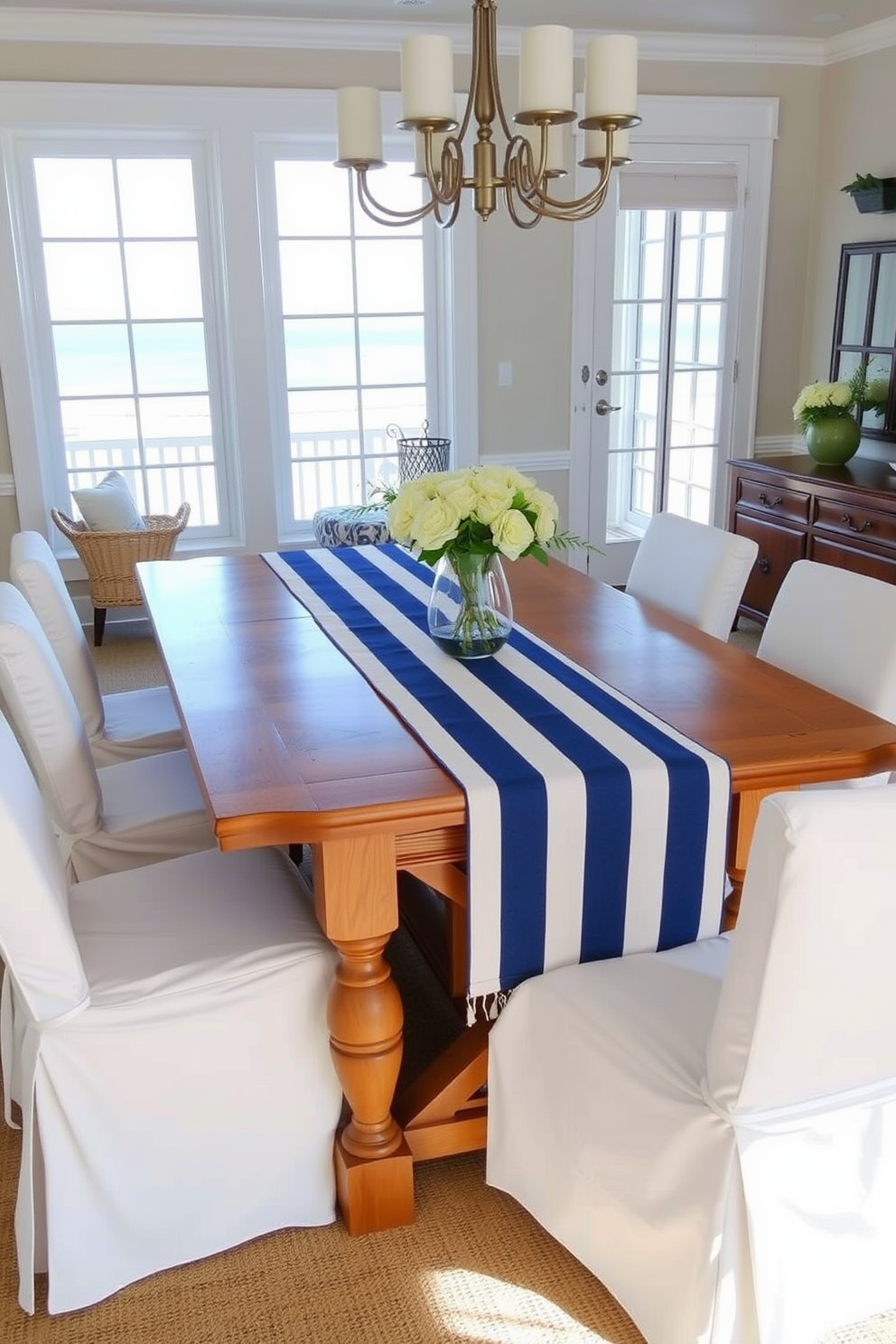 A beach dining room featuring a nautical theme with a blue and white striped table runner elegantly draped across a wooden dining table. Surrounding the table are white slipcovered chairs that enhance the coastal vibe, while large windows allow natural light to flood the space.