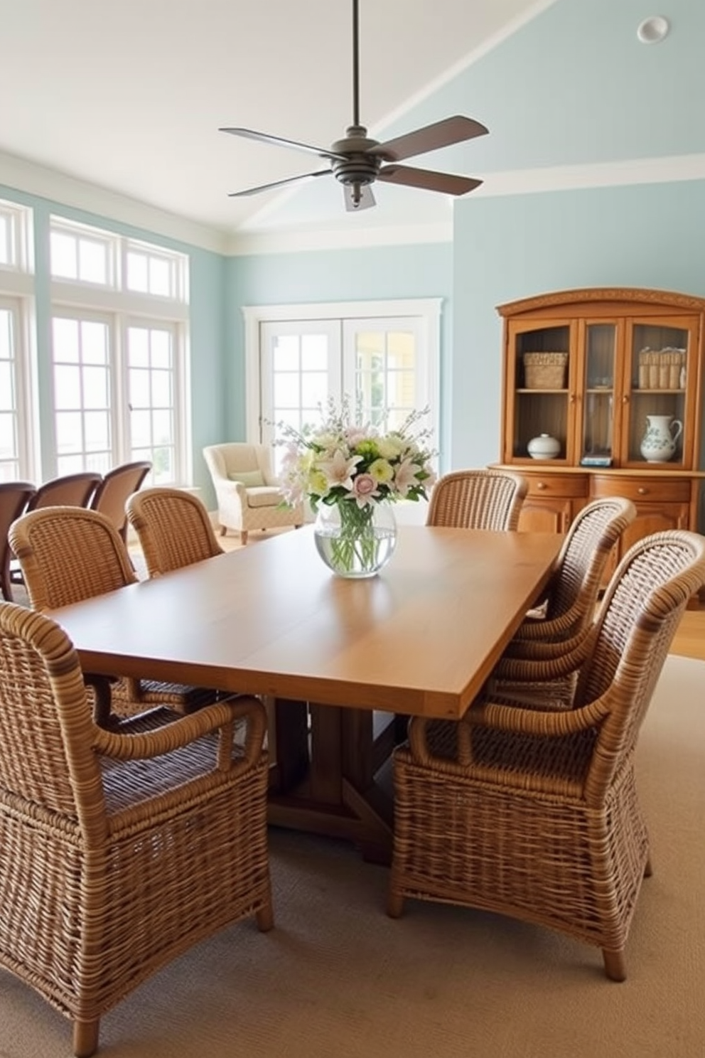 Woven rattan chairs are arranged around a large wooden dining table, creating a warm and inviting atmosphere. The room features large windows that allow natural light to flood in, highlighting the coastal decor and ocean-inspired color palette. Light blue walls complement the sandy beige tones of the table and chairs. A centerpiece of fresh flowers in a glass vase adds a touch of elegance to the beach-themed dining space.