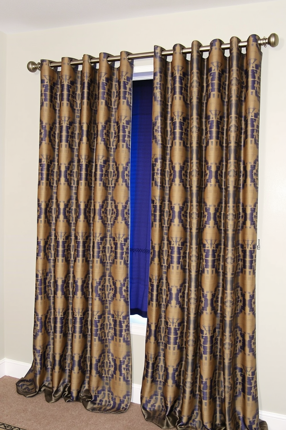 A pair of patterned blackout curtains drapes elegantly over a large window, providing both style and functionality. The fabric features a modern geometric design in shades of navy and gold, complementing the room's decor while blocking out light for a restful atmosphere. The curtains are hung from a sleek, brushed nickel rod that adds a contemporary touch to the overall look. Soft folds cascade to the floor, creating a luxurious feel and enhancing the cozy ambiance of the bedroom.