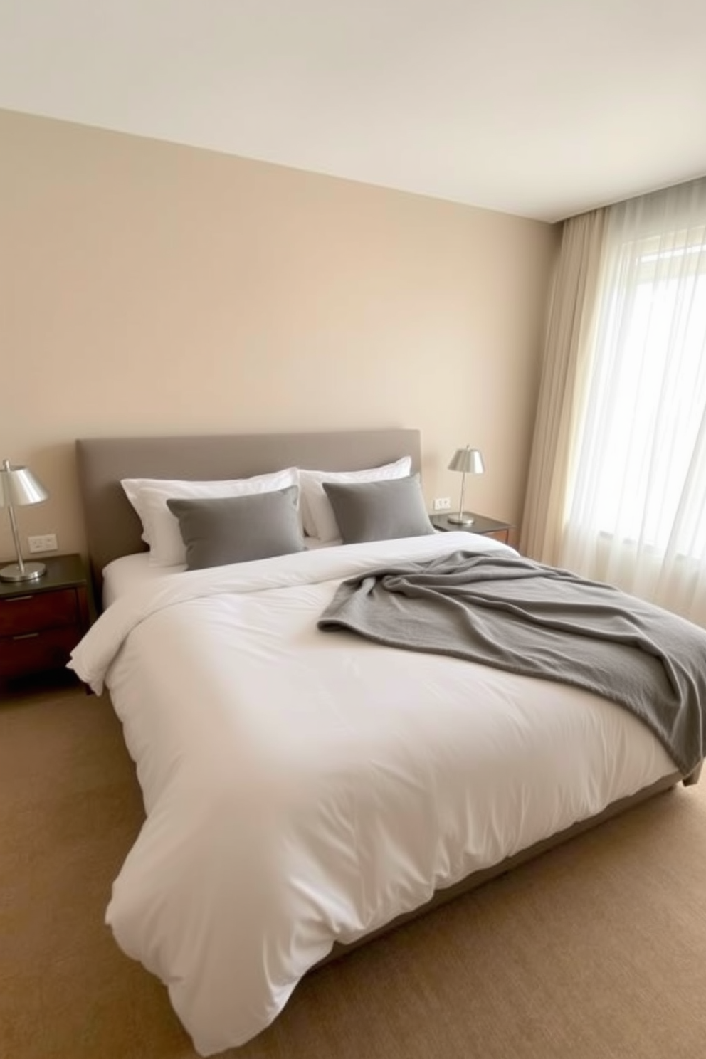 A serene bedroom featuring a plush king-sized bed with crisp white linens and a soft gray throw blanket. The walls are painted in a light beige hue, creating a warm and inviting atmosphere. On either side of the bed, there are sleek nightstands with minimalist lamps providing soft lighting. A large window with sheer curtains allows natural light to filter in, illuminating the space beautifully.