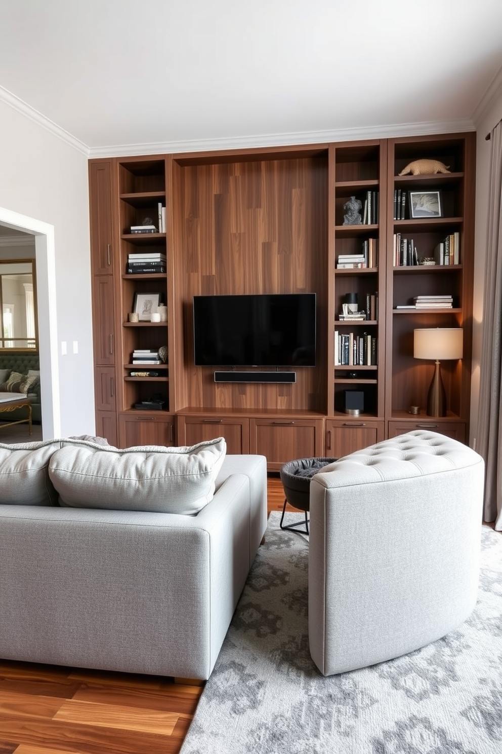 A cozy living room that seamlessly integrates a compact entertainment center. The space features a plush sectional sofa in a soft gray fabric, positioned to face a sleek wall-mounted TV surrounded by built-in shelves filled with books and decorative items. The bedroom area is defined by a stylish area rug that adds warmth to the hardwood floor. A queen-sized bed with a tufted headboard sits against the wall, flanked by modern nightstands with lamps that complement the overall aesthetic.