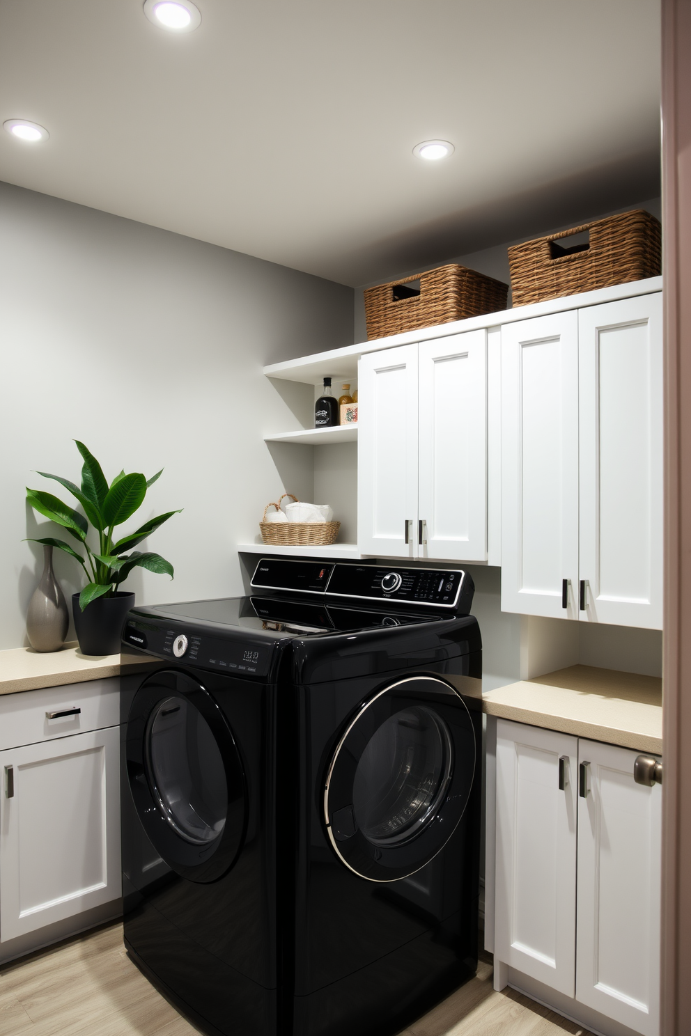 A modern laundry room featuring sleek black appliances that create a bold contrast against white cabinetry. The space is illuminated by recessed lighting, highlighting the organized layout and functional design. The walls are painted in a soft gray hue, providing a subtle backdrop for the striking black washer and dryer. A stylish countertop extends above the appliances, adorned with decorative baskets for storage and a potted plant for a touch of greenery.