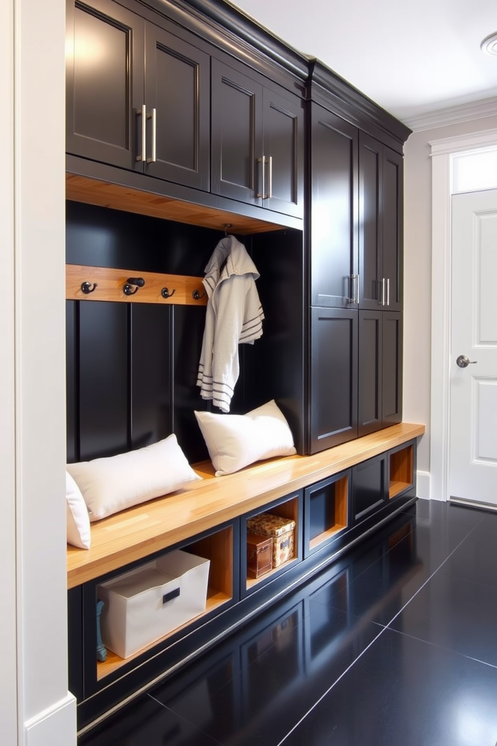 A stylish mudroom featuring a combination of black cabinetry and natural wood accents. The space includes built-in benches with plush cushions, hooks for coats, and a sleek black tile floor that contrasts beautifully with the warm wood tones.