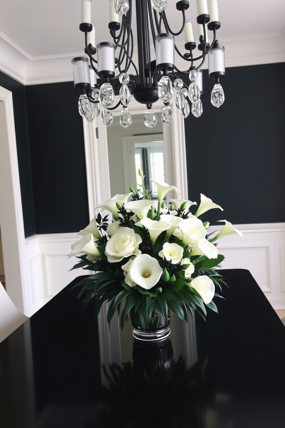 A stunning black and white floral centerpiece arrangement sits elegantly on a sleek black dining table. The arrangement features a mix of large white peonies and delicate black calla lilies, complemented by lush greenery. The dining room is designed with a modern aesthetic, featuring black accent walls and white wainscoting. A statement chandelier with black and white elements hangs above the table, creating a striking focal point in the space.