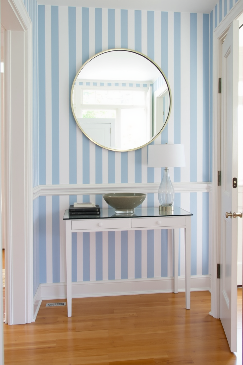 A bright and airy foyer features blue and white striped wallpaper that creates a refreshing atmosphere. The space is adorned with a sleek console table, topped with a decorative bowl and a stylish lamp. The flooring is a light hardwood that complements the wallpaper beautifully. A large round mirror hangs above the console, reflecting natural light from a nearby window.