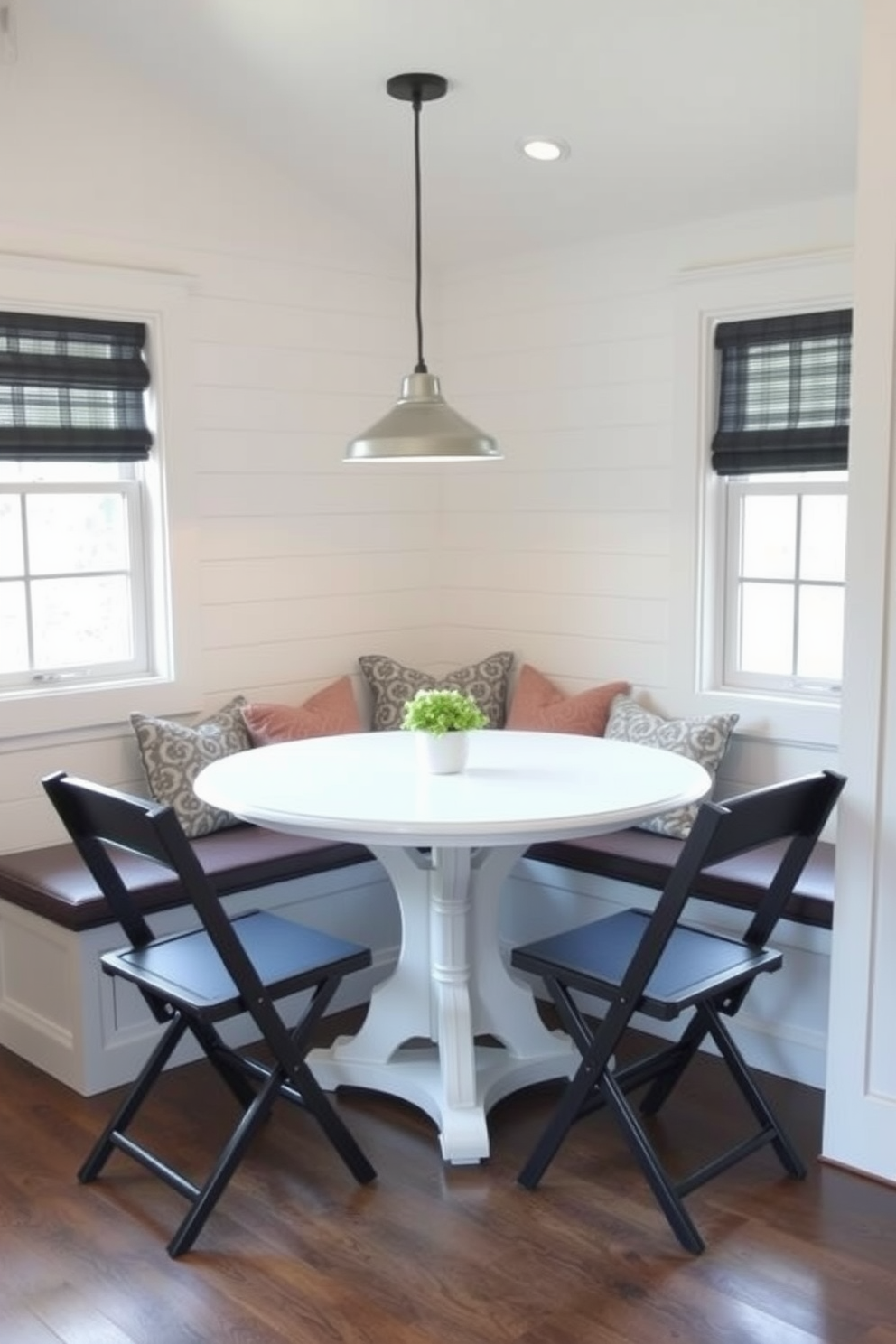 A cozy breakfast nook featuring a round table surrounded by foldable chairs that can be tucked away when not in use. The walls are adorned with light-colored shiplap and a large window allows natural light to flood the space, enhancing the inviting atmosphere. In one corner, a built-in bench with cushions provides extra seating and storage underneath. A small pendant light hangs above the table, creating a warm ambiance perfect for morning coffee and casual meals.