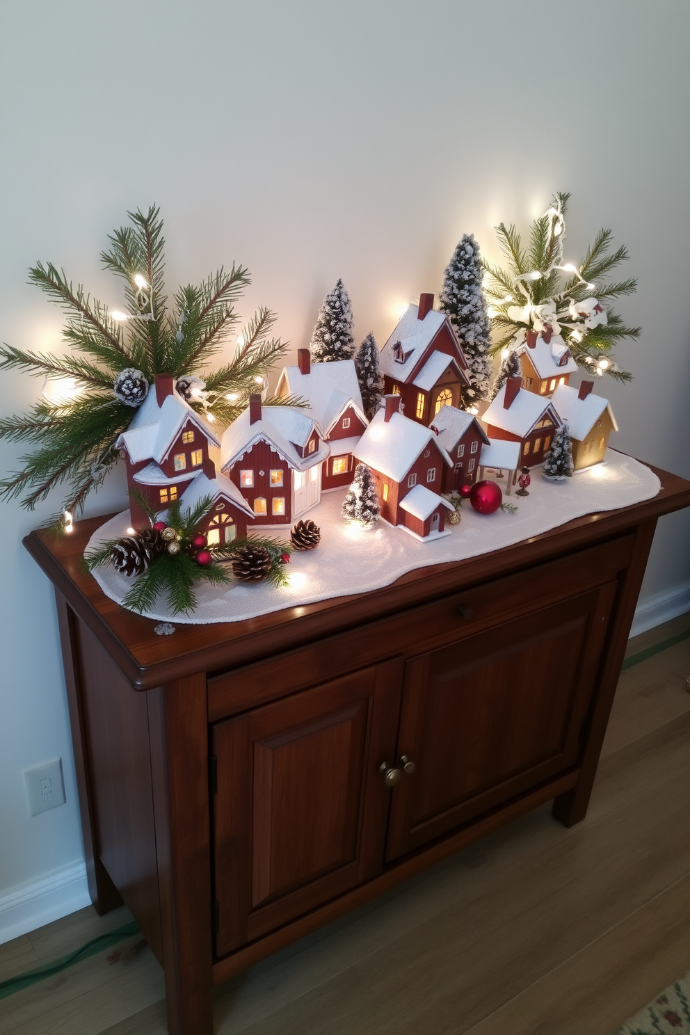 A cozy winter village scene is displayed on a wooden console table, featuring a collection of charming miniature houses dusted with snow. Soft white lights twinkle around the village, creating a warm and inviting atmosphere for the holiday season. Sprigs of evergreen and pinecones are artfully arranged among the houses, adding a natural touch to the festive decor. A few decorative ornaments in red and gold hues are scattered throughout, enhancing the Christmas spirit.