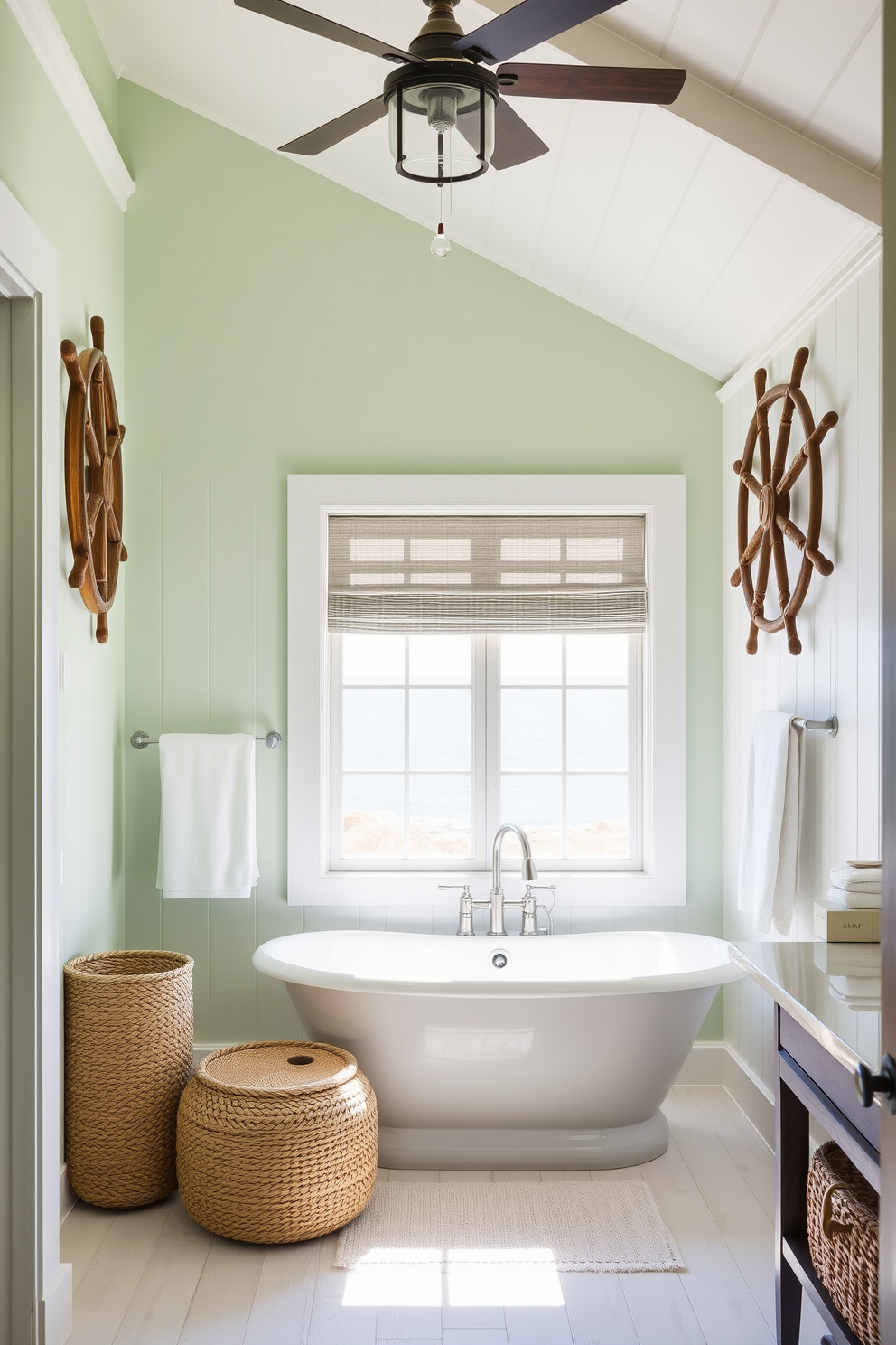 Antique ship wheels are prominently displayed as wall art, adding a nautical charm to the space. The walls are painted in a soft seafoam green, complementing the white shiplap paneling. The bathroom features a freestanding soaking tub positioned beneath a large window, offering a view of the ocean. Natural light floods the room, enhancing the airy feel created by light wood accents and woven baskets for storage.