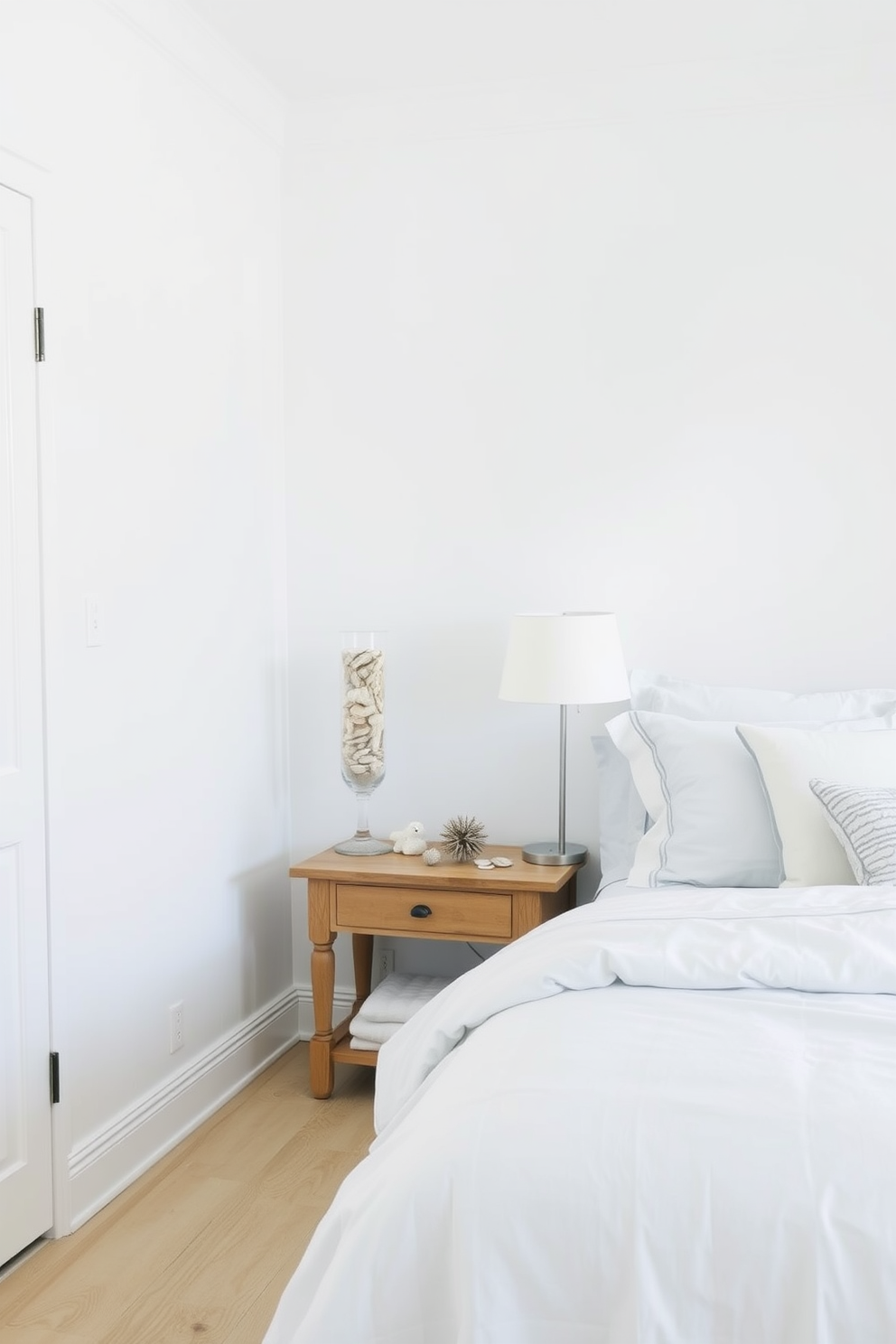 A serene coastal bedroom setting. The room features soft white walls and light wood flooring, creating a bright and airy atmosphere. In the corner, glass vases filled with sand and shells are artfully arranged on a wooden side table. The bedding is a mix of light blue and white linens, complementing the coastal theme with subtle nautical patterns.