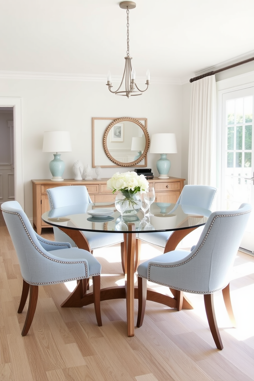 A glass dining table with elegant wooden legs sits at the center of a bright coastal dining room. Surrounding the table are light blue upholstered chairs that complement the airy, beach-inspired decor.