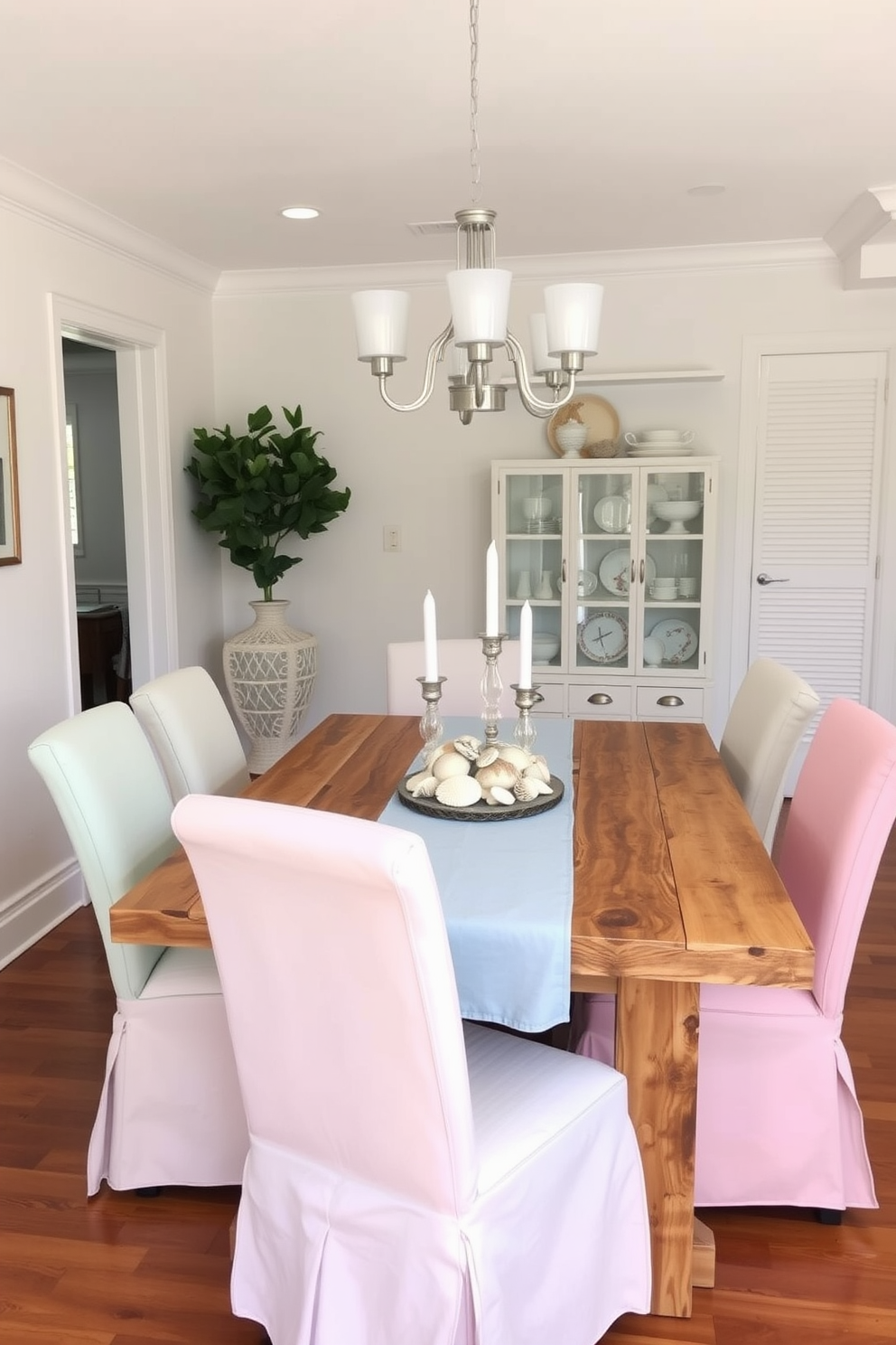 A coastal dining room featuring simple slipcovered chairs in soft pastel colors. The table is made of reclaimed wood and is adorned with a light blue table runner and a centerpiece of seashells and candles.