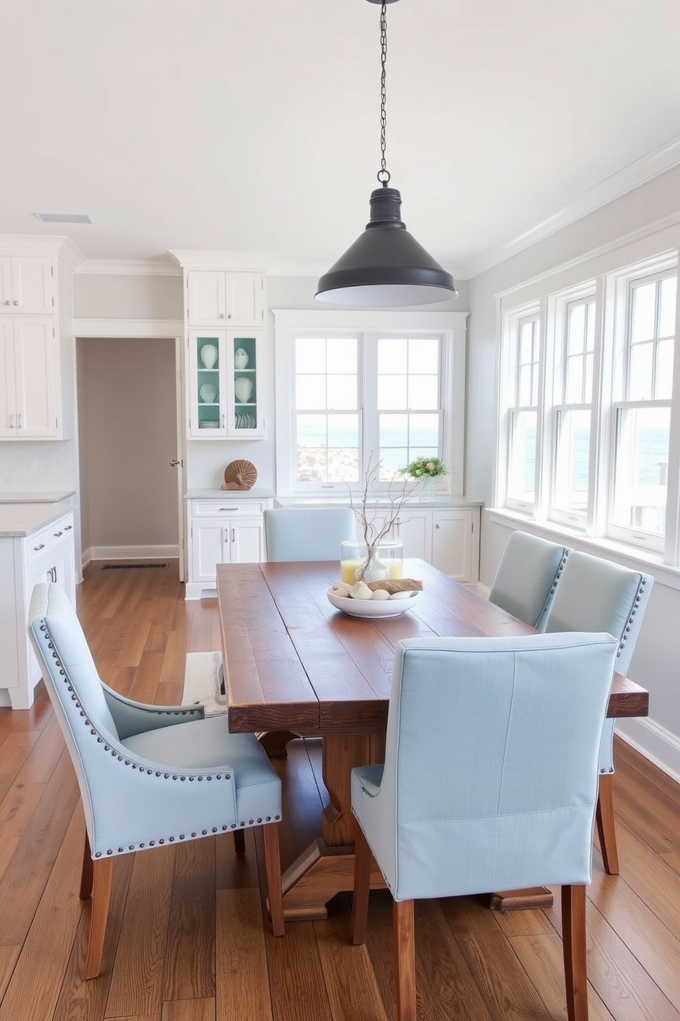 Bright white cabinetry creates a clean and airy atmosphere in the coastal dining room. The dining table is made of reclaimed wood, surrounded by light blue upholstered chairs that add a touch of color. Large windows allow natural light to flood the space, enhancing the bright and inviting feel. Nautical decor elements, such as seashells and driftwood accents, complement the overall coastal theme.