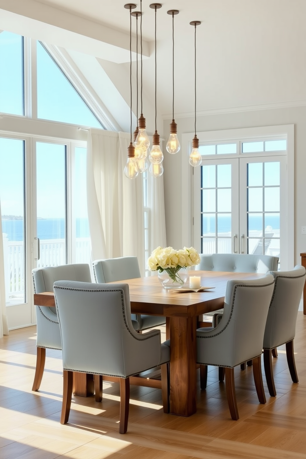 A coastal dining room with a large wooden table surrounded by light blue upholstered chairs. The space is illuminated by hanging pendant lights with glass bulbs that create a warm and inviting atmosphere. Large windows allow natural light to flood in, showcasing a view of the ocean. Soft white curtains frame the windows, adding a touch of elegance to the beach-inspired decor.