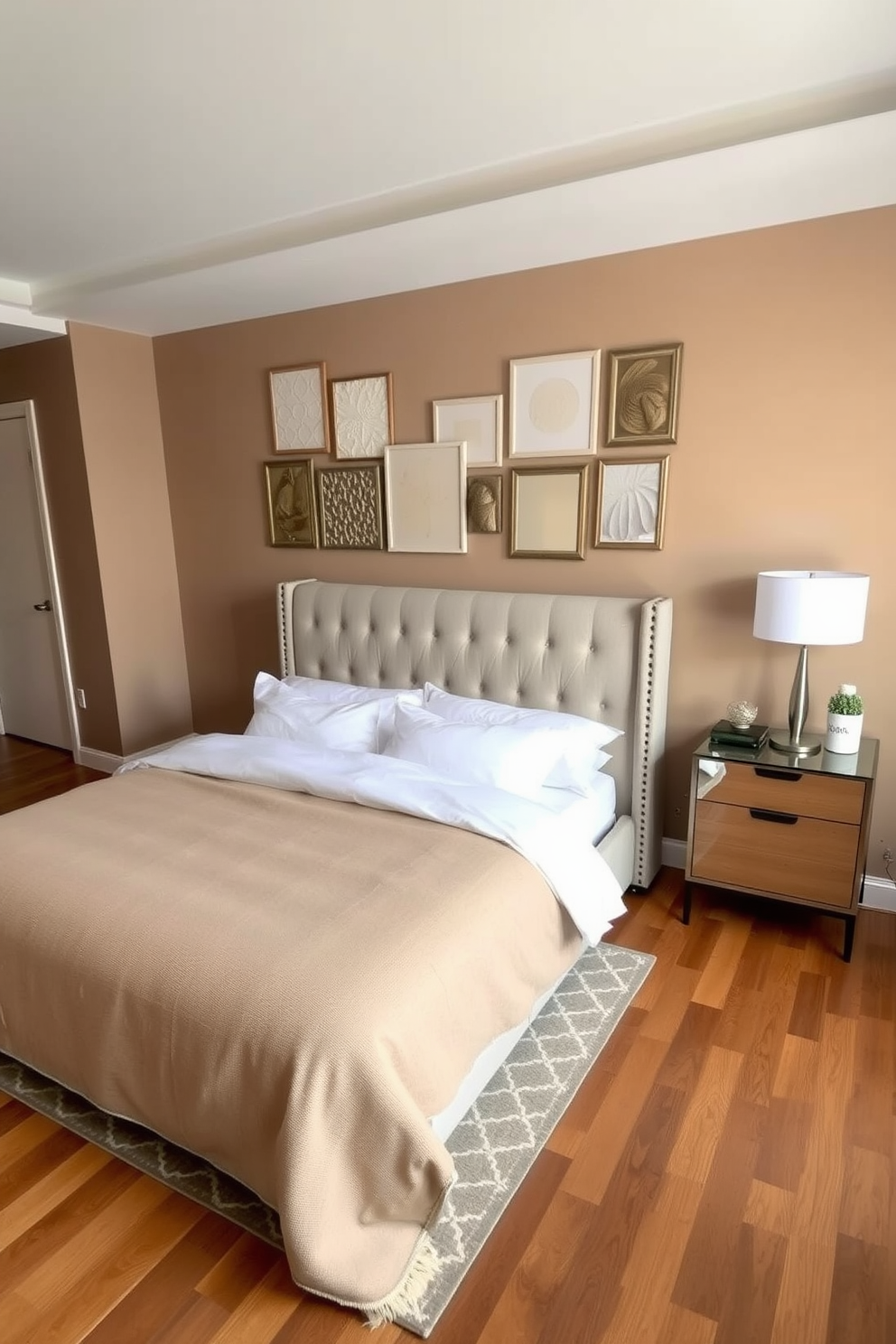 A cozy condo bedroom featuring a plush upholstered bed with a tufted headboard dressed in soft white linens. The walls are painted a warm taupe, and a large area rug with a geometric pattern adds warmth to the hardwood floor. In one corner, a sleek nightstand with a modern lamp complements the bed. A gallery wall of framed artwork in various textures and finishes adds personality and depth to the space.