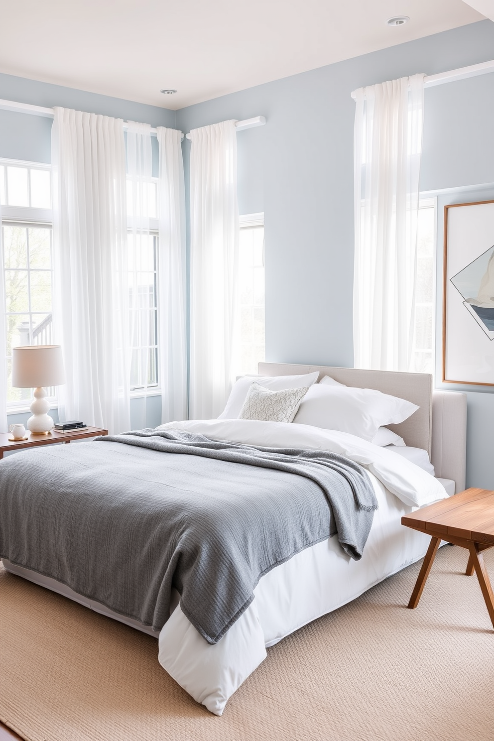 A serene bedroom retreat featuring a plush king-sized bed dressed in soft white linens and an oversized gray throw blanket. Large windows allow natural light to flood the space, framed by sheer white curtains that gently sway in the breeze. On one side of the bed, a sleek nightstand holds a modern lamp with a warm glow, while on the opposite side, a minimalist wooden bench adds functionality. The walls are painted in a calming light blue hue, complemented by abstract artwork that adds a touch of elegance.