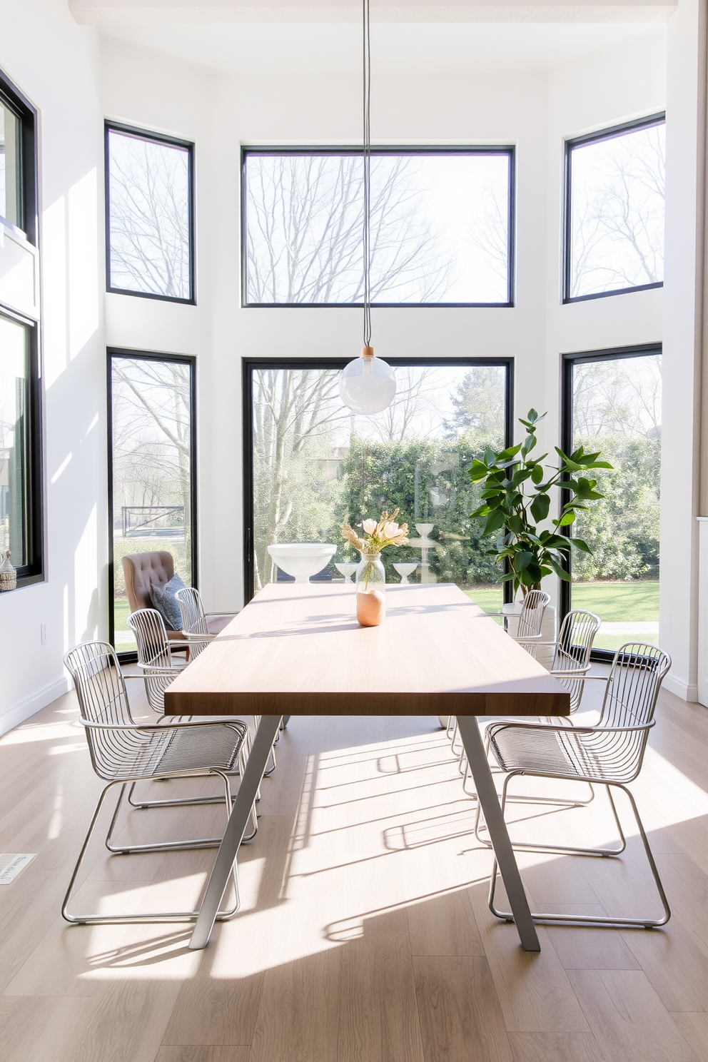 A minimalist dining table is set with simple white dinnerware and sleek stainless steel cutlery. A single green plant in a small ceramic pot serves as the centerpiece, adding a touch of nature to the clean aesthetic. The dining room features a light wood table surrounded by clear acrylic chairs that create an open and airy feel. Soft natural light floods the space through large windows, enhancing the contemporary design with a fresh and inviting atmosphere.