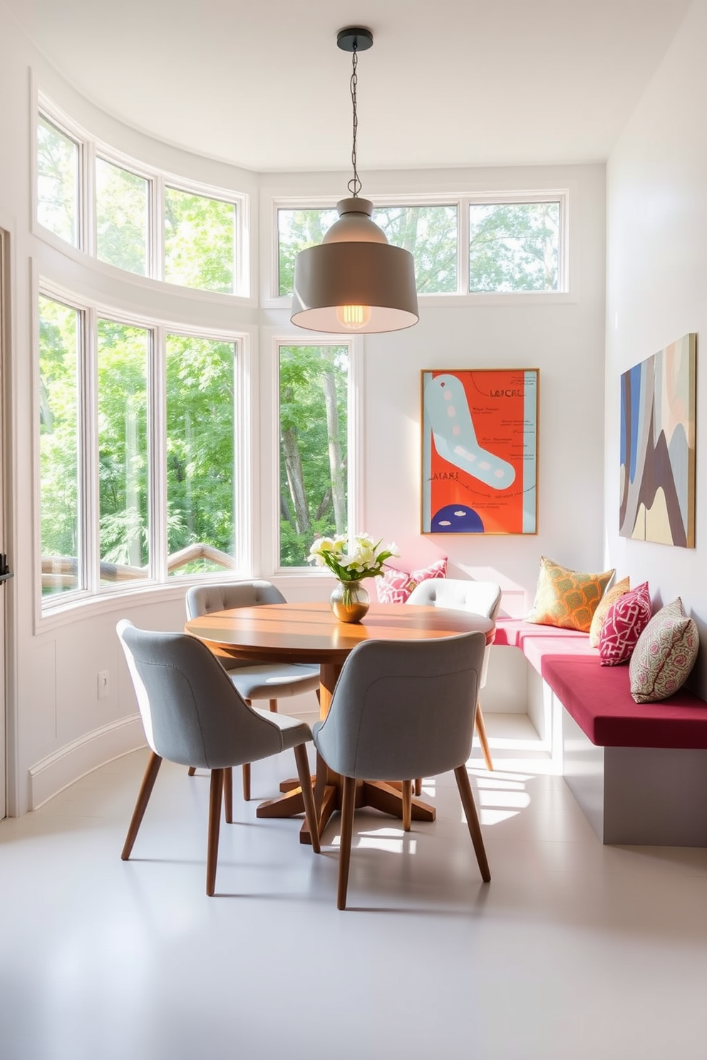 A contemporary dining nook features a round wooden table surrounded by upholstered chairs in soft gray. Large windows allow natural light to flood the space, complemented by a statement pendant light hanging above the table. The walls are painted in a crisp white, creating a clean backdrop for vibrant artwork. A built-in bench with colorful cushions lines one wall, providing additional seating and a cozy atmosphere.