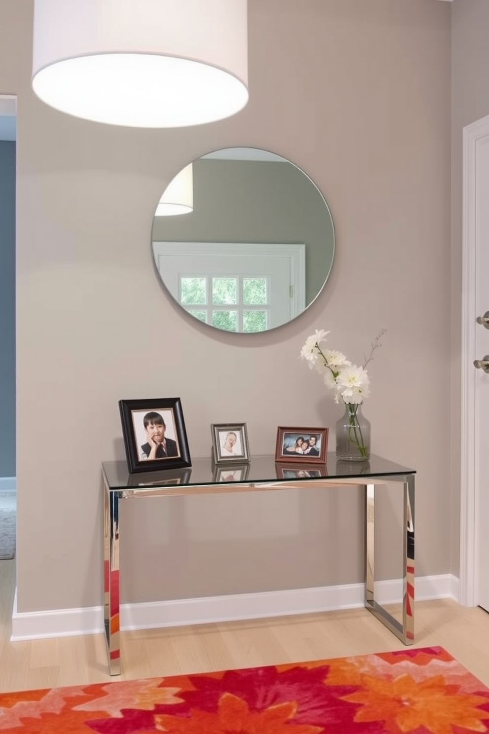 A contemporary foyer features a sleek console table made of glass and metal. Above the table, a large round mirror reflects the light from a stylish pendant lamp. Framed family photos are artfully arranged on the console table, adding warmth and a personal touch. The walls are painted in a soft gray, complemented by a vibrant area rug that anchors the space.