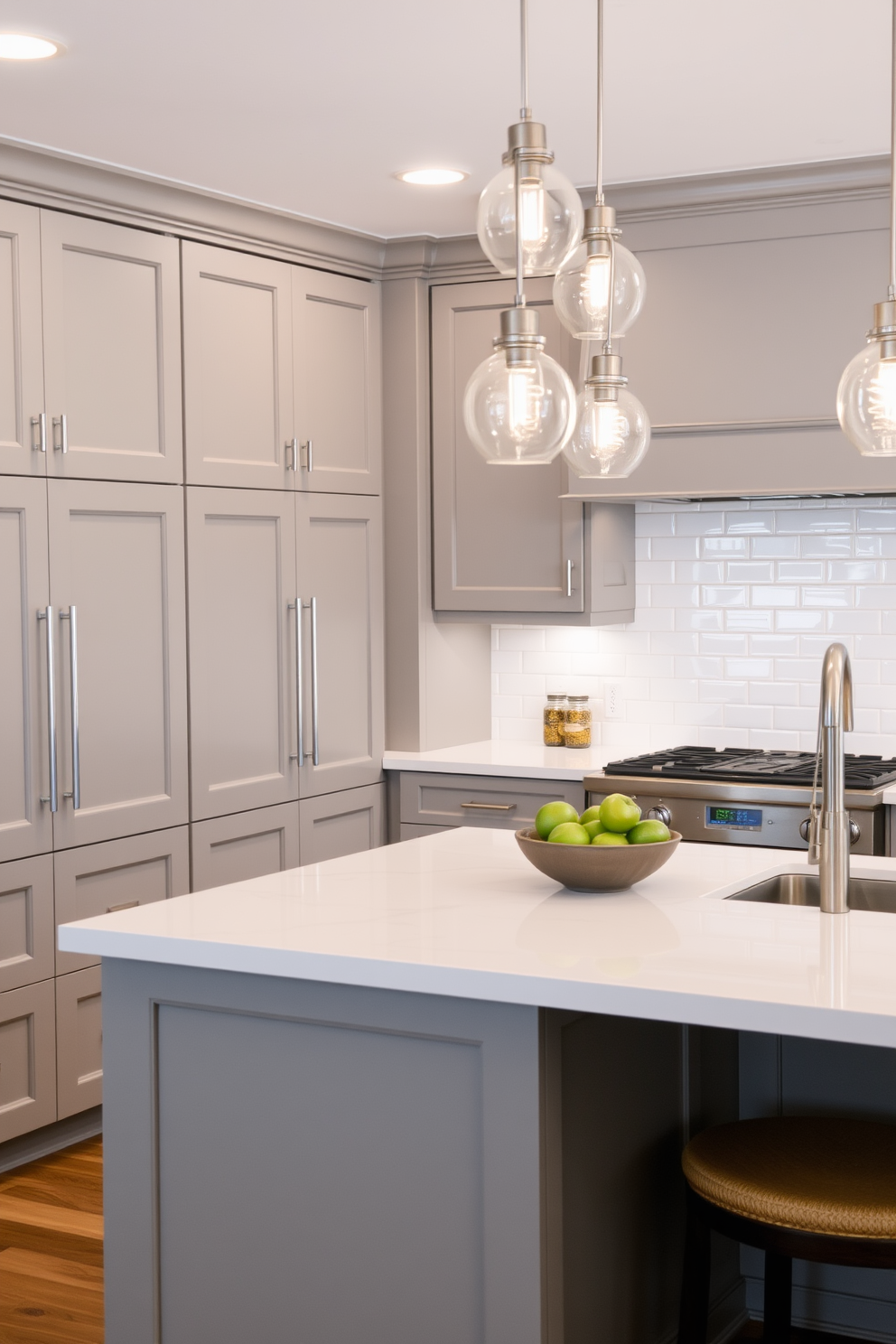 A contemporary kitchen featuring sleek cabinetry in a soft gray finish with brushed nickel hardware. The backsplash is adorned with glossy white subway tiles that create a timeless and clean look. An island with a white quartz countertop provides ample space for meal preparation and casual dining. Pendant lights with a modern design hang above the island, adding a touch of elegance to the overall aesthetic.