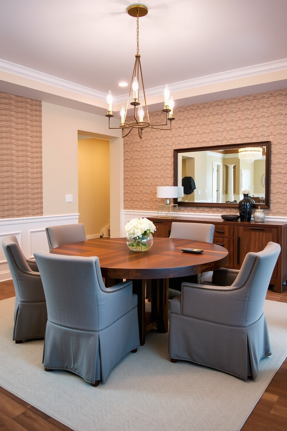 A stylish dining area features a large round table made of reclaimed wood surrounded by upholstered chairs in soft gray fabric. Above the table, a modern chandelier with gold accents hangs, providing warm ambient lighting for gatherings. The walls are painted in a soft beige tone, complemented by a textured wallpaper on one accent wall. A sideboard in a rich walnut finish sits against the wall, adorned with decorative items and a large mirror that reflects the inviting atmosphere of the space.