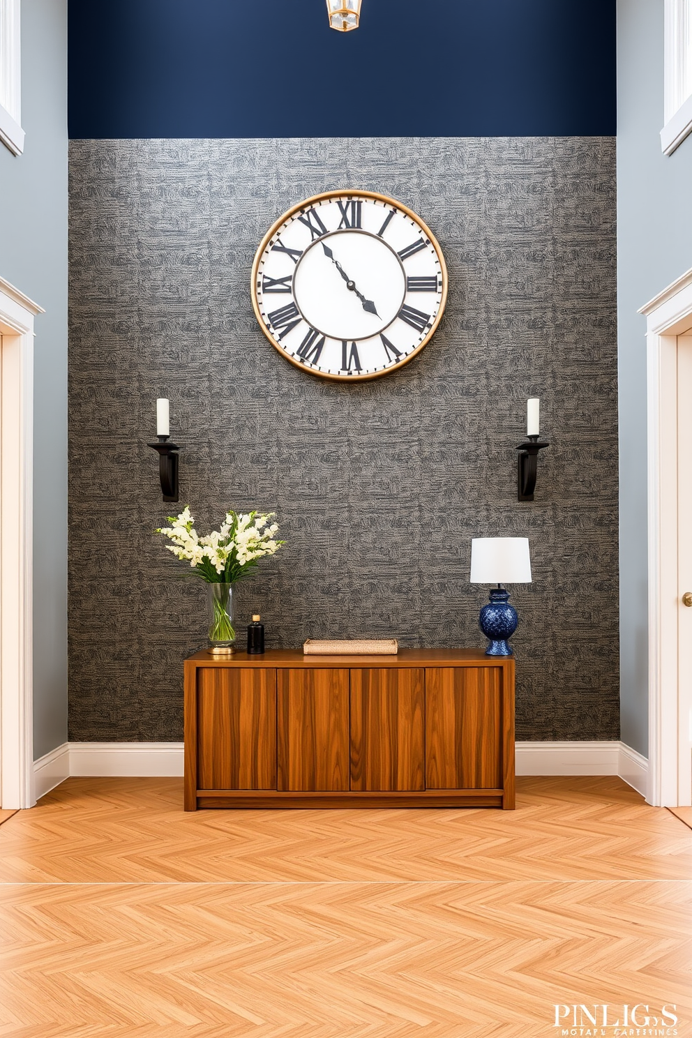 An elegant console table is positioned against the wall in the entryway. It is adorned with a stylish lamp, a decorative bowl, and a few carefully arranged books. The wall behind the console table features a subtle textured wallpaper in a soft neutral tone. Above the table, a large framed mirror reflects the warm light from the lamp, enhancing the space's inviting atmosphere.