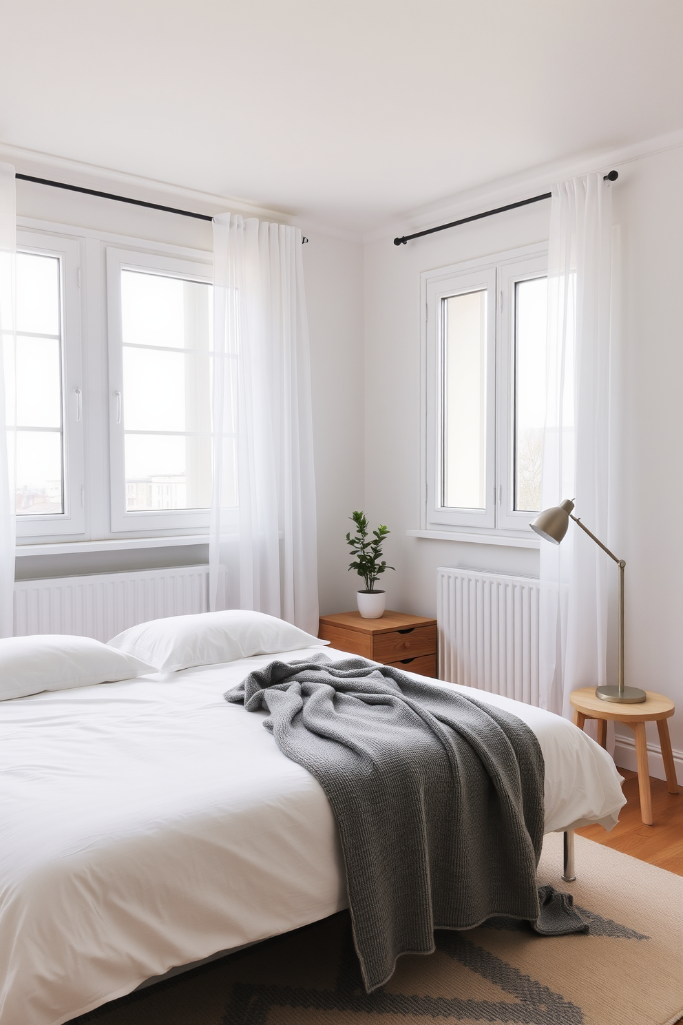 A serene European bedroom featuring minimalist decor with a neutral color palette. The room includes a low-profile bed with crisp white linens and a soft gray throw blanket draped casually across the foot. Natural light floods the space through large windows adorned with sheer curtains. A simple wooden nightstand holds a small potted plant and a sleek lamp, enhancing the clean aesthetic.