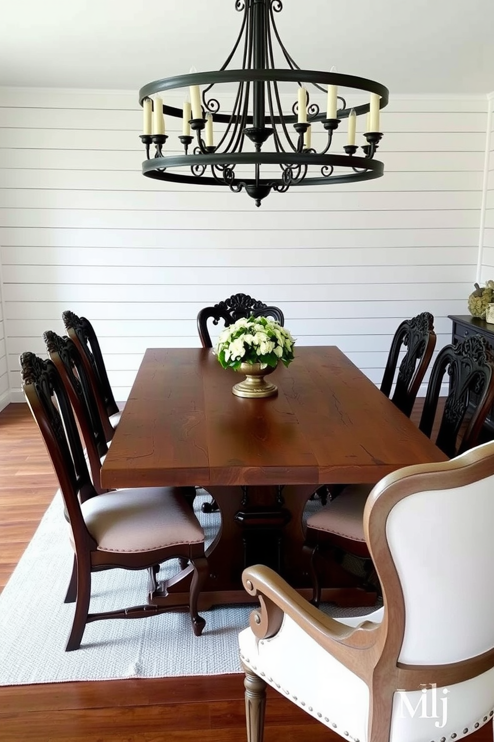 A rustic farmhouse table made of reclaimed wood is the centerpiece of the dining room. Surrounding the table are vintage chairs with intricate carvings and upholstered seats in a soft neutral fabric. The walls are adorned with shiplap paneling painted in a warm white hue. A large, wrought iron chandelier hangs above the table, casting a warm glow over the space.