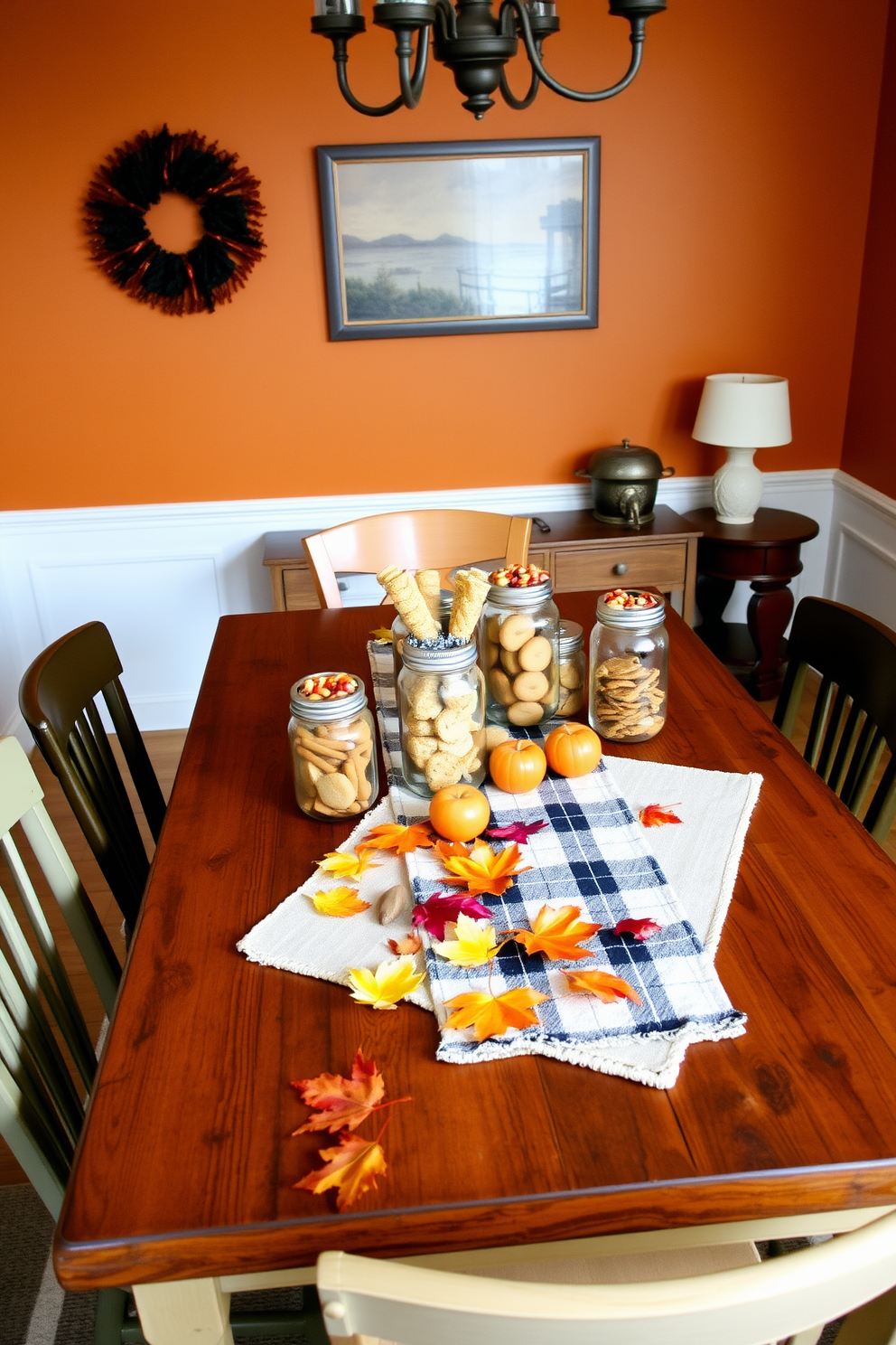 A cozy fall dining room setting. A rustic wooden table is adorned with mason jars filled with seasonal treats like pumpkin spice cookies and caramel apples. Surrounding the table are mismatched chairs in warm tones, and a soft plaid table runner adds texture. The walls are painted in a warm amber hue, and autumn leaves are scattered decoratively across the table.