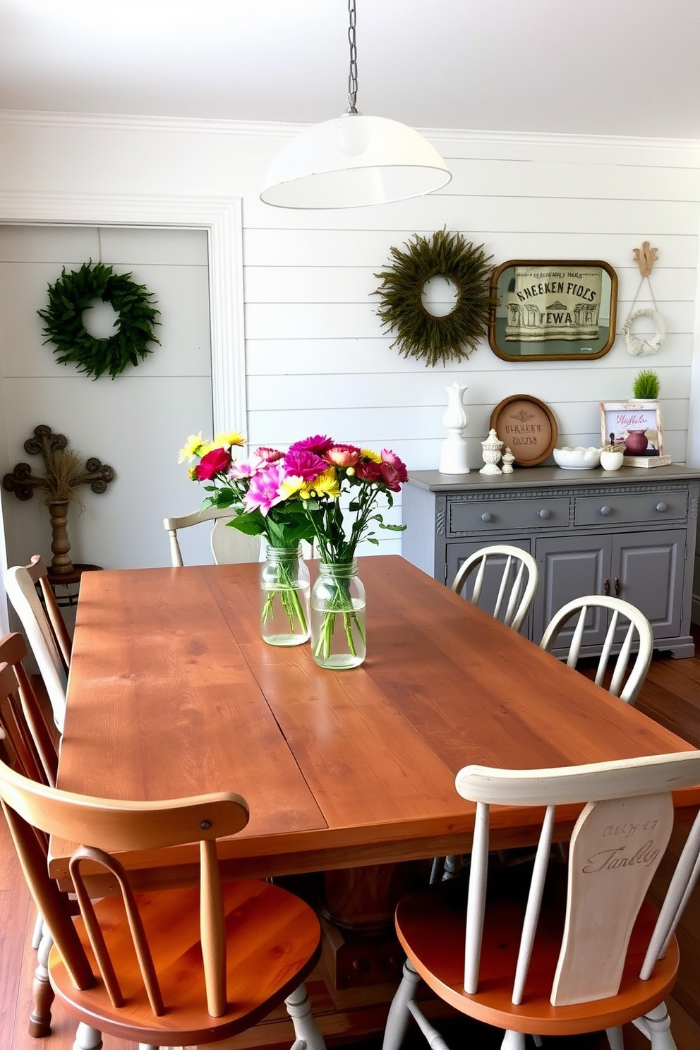 A charming farmhouse dining room featuring a rustic wooden table surrounded by mismatched chairs. Fresh flowers in mason jar vases are placed at the center of the table, adding a vibrant touch to the cozy atmosphere. The walls are adorned with shiplap and painted in a soft white hue, creating a bright and airy feel. A vintage sideboard holds additional decor items and provides storage for dining essentials.