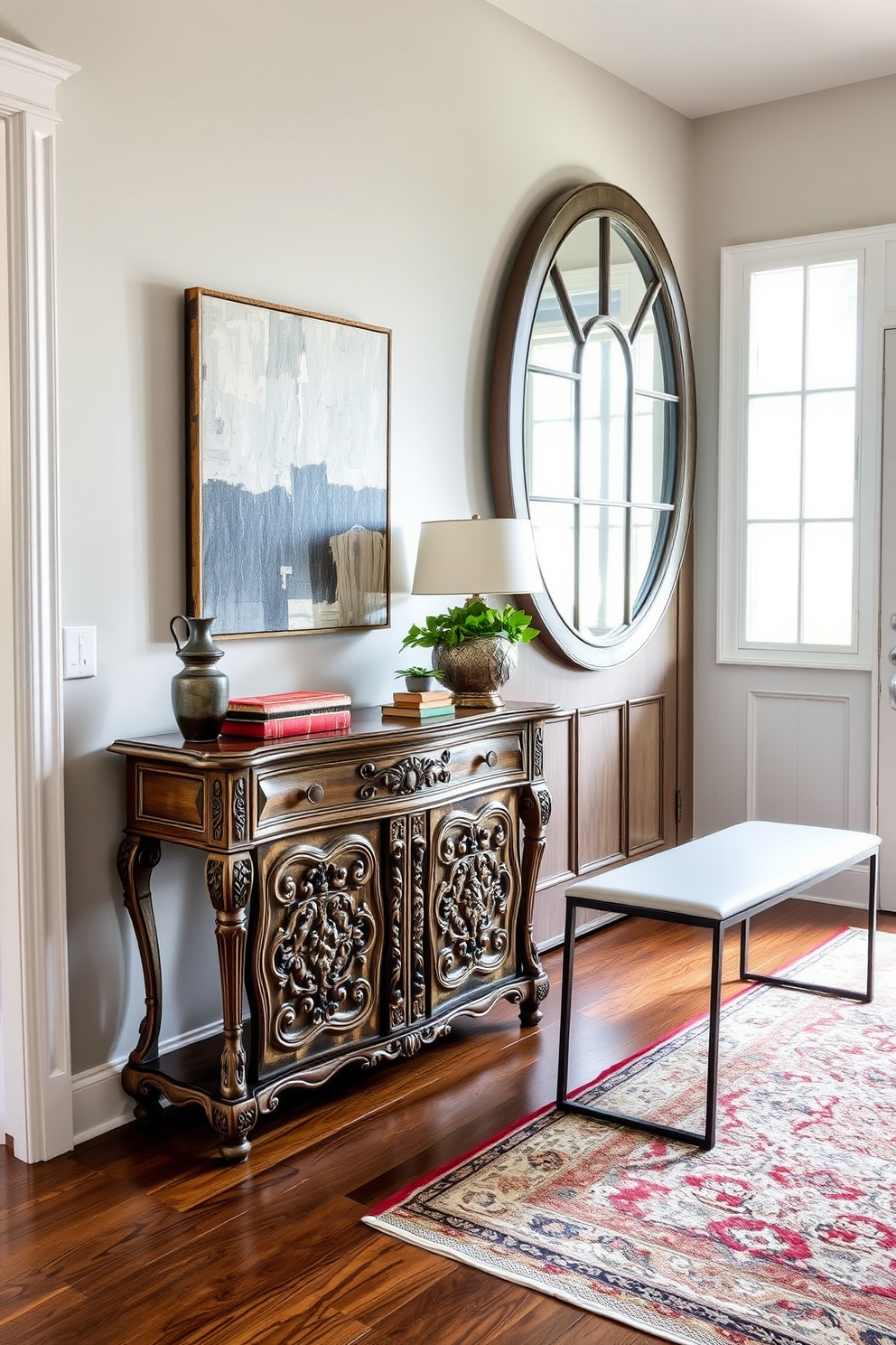 A welcoming foyer with a blend of vintage and modern decor elements. An antique console table with intricate carvings stands against the wall, adorned with a contemporary abstract painting above it. To the right, a sleek modern bench with clean lines offers seating, paired with a vintage area rug that adds warmth. A large round mirror with a distressed frame reflects the natural light coming from a nearby window, enhancing the inviting atmosphere.