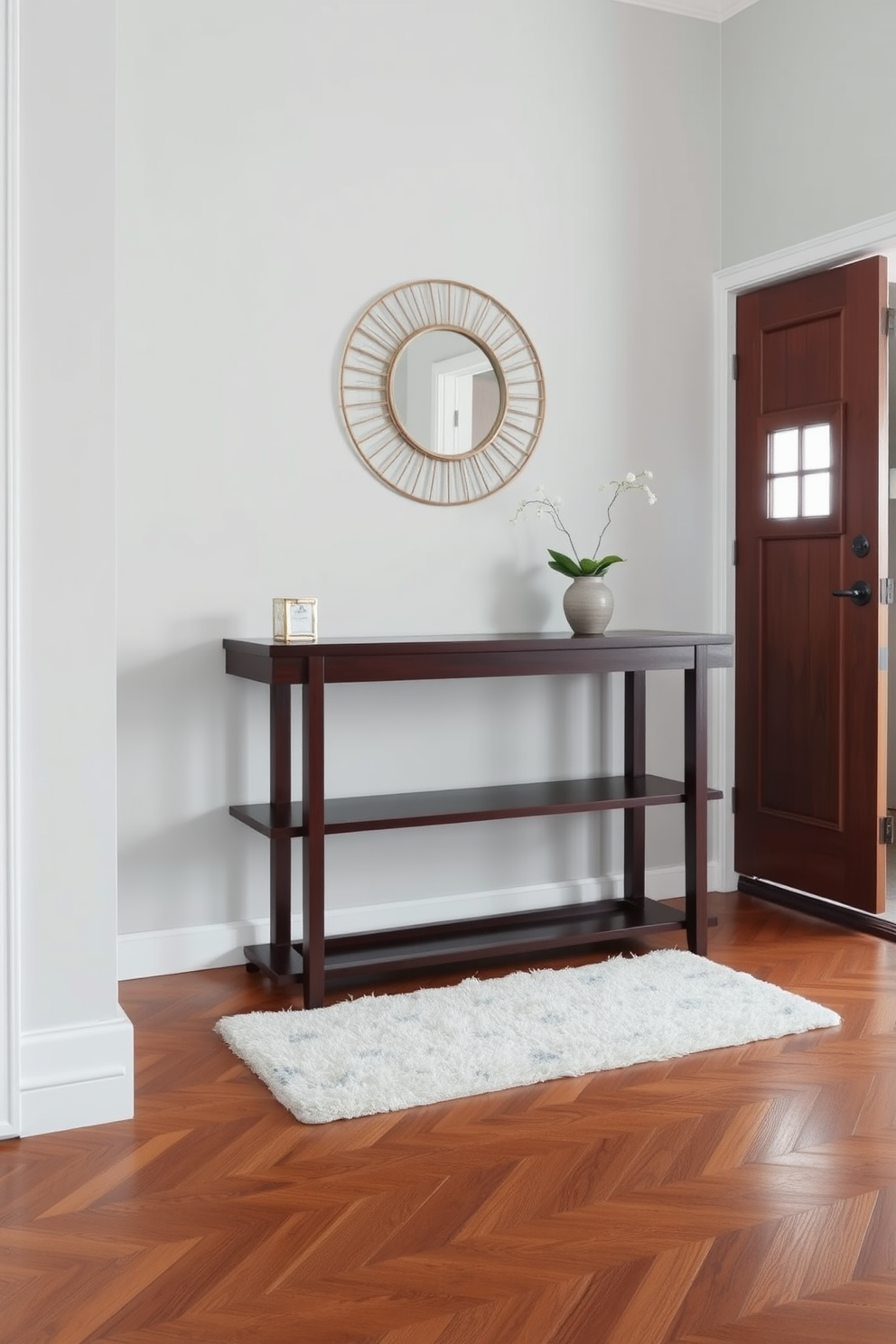 A stylish foyer featuring a sleek console table made of dark wood against a soft gray wall. The table is adorned with a decorative mirror above it and a small potted plant on one side, creating an inviting entrance. The floor is finished with elegant herringbone-patterned hardwood, complementing the overall aesthetic. A plush area rug lies beneath the console table, adding warmth and texture to the space.