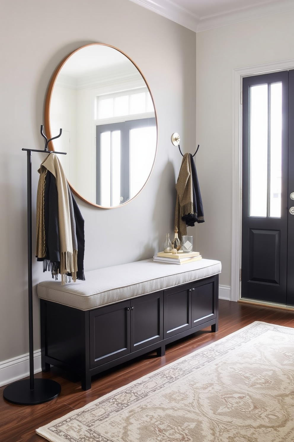 A chic bench with storage underneath is positioned against the wall in the foyer. The bench is upholstered in a soft gray fabric, and above it, a large round mirror reflects natural light coming through the nearby window. On one side of the bench, a stylish coat rack holds a few elegant jackets and scarves. The floor is adorned with a patterned area rug that adds warmth and character to the entryway space.