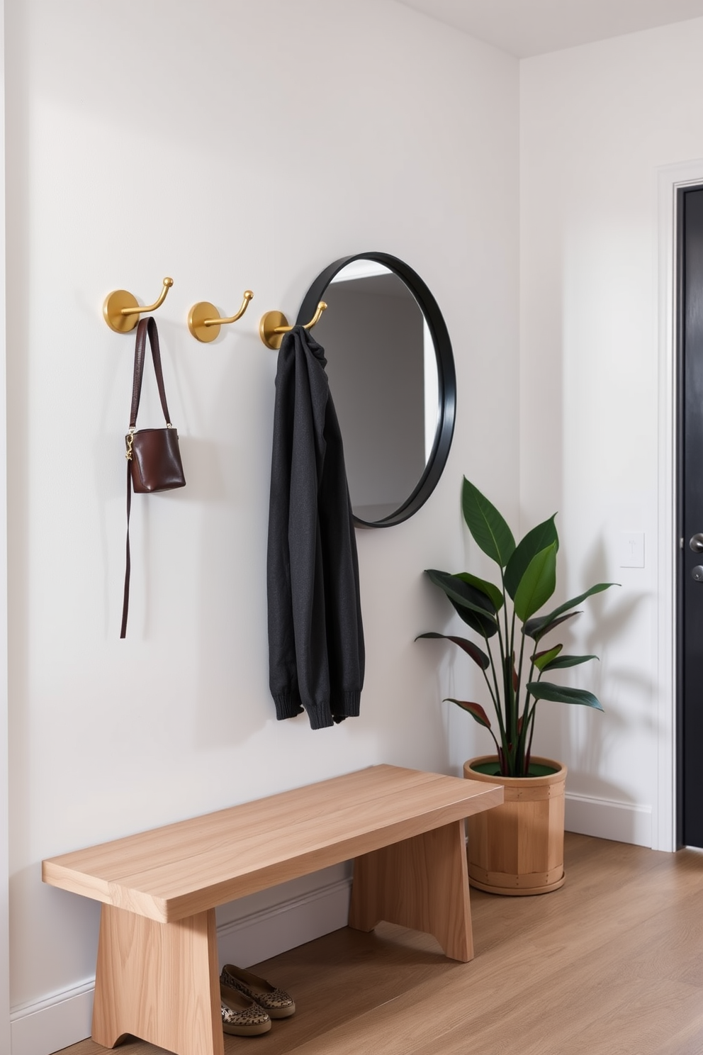 Stylish wall hooks made of brushed brass are mounted on a sleek white wall, providing easy access for coats and bags. Below, a minimalist bench with a light oak finish offers a convenient spot to sit while putting on shoes. The foyer features a large round mirror framed in black, reflecting natural light and creating an inviting atmosphere. A potted plant sits in the corner, adding a touch of greenery and warmth to the entryway.