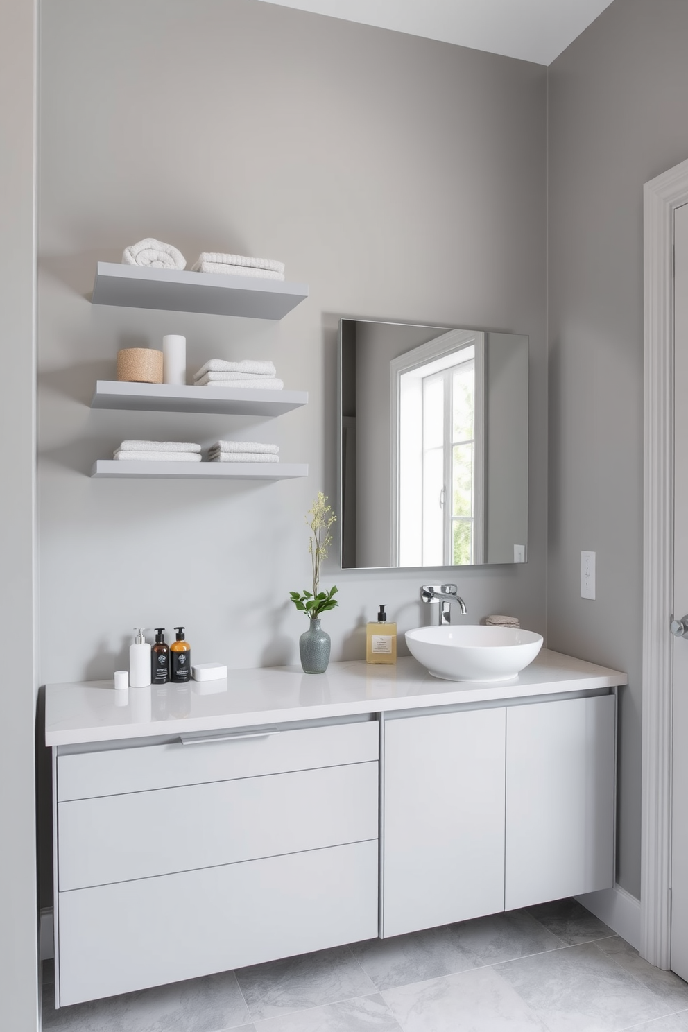 A contemporary gray bathroom features sleek storage solutions that blend functionality with style. Floating shelves in a light gray finish are mounted above a modern vanity, providing ample space for toiletries and decorative items. The cabinetry is designed with clean lines and a minimalist aesthetic, offering a seamless look. Soft gray tiles cover the floor, complemented by a large mirror that reflects natural light, enhancing the room's spacious feel.