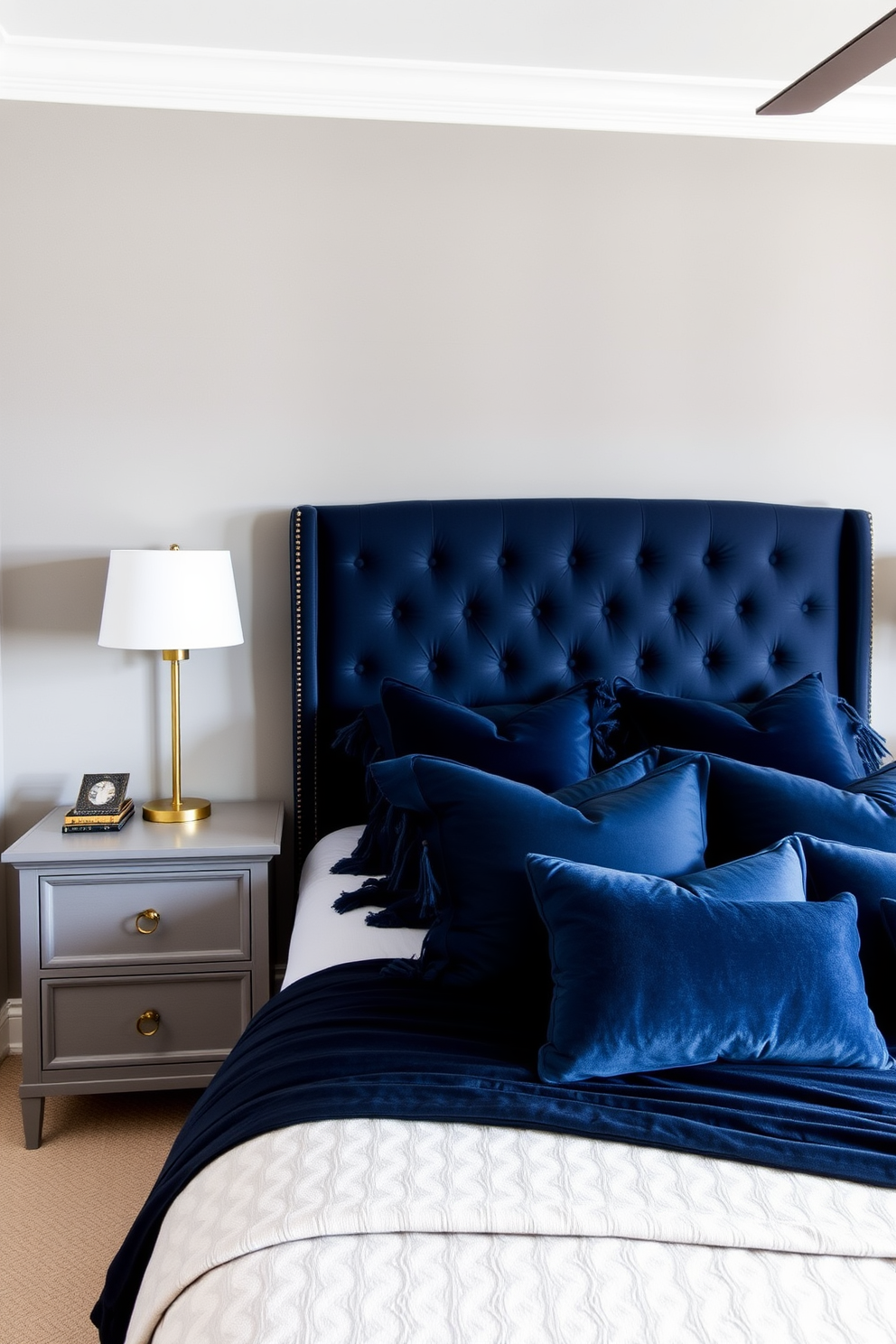 A cozy bedroom featuring a classic gray and navy blue palette. The walls are painted in a soft gray, while the bedding incorporates rich navy blue accents for a sophisticated touch. A plush navy blue upholstered headboard serves as the focal point of the room. Complementing the headboard, gray nightstands with elegant brass hardware sit on either side of a stylish bed.