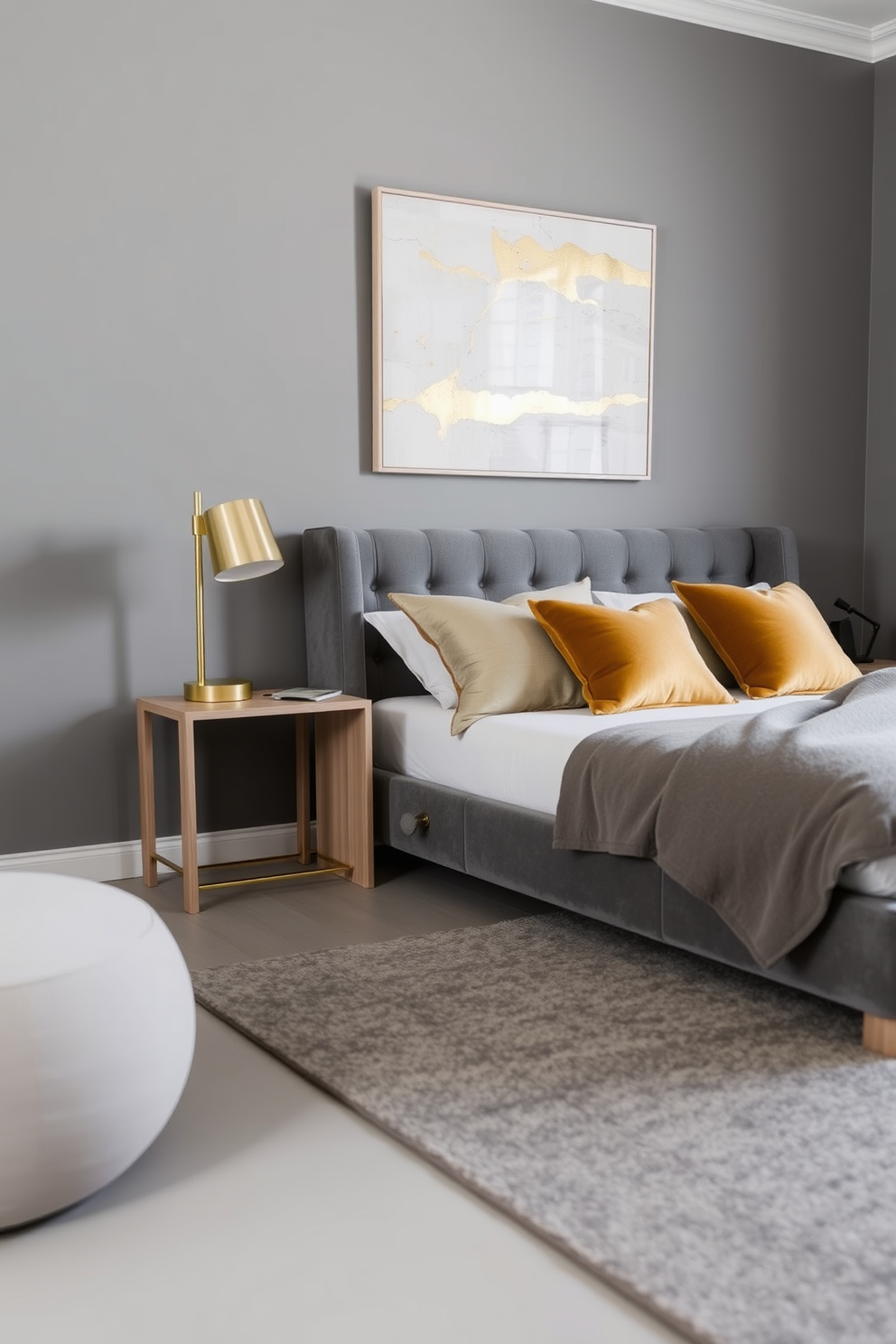 A serene bedroom setting featuring muted gray tones throughout the space. The walls are painted in a soft gray shade, complemented by a plush gray upholstered bed adorned with golden accent pillows. A stylish nightstand made of light wood sits beside the bed, topped with a sleek golden lamp. The floor is covered with a cozy gray area rug, and a piece of abstract art with hints of gold hangs above the bed.