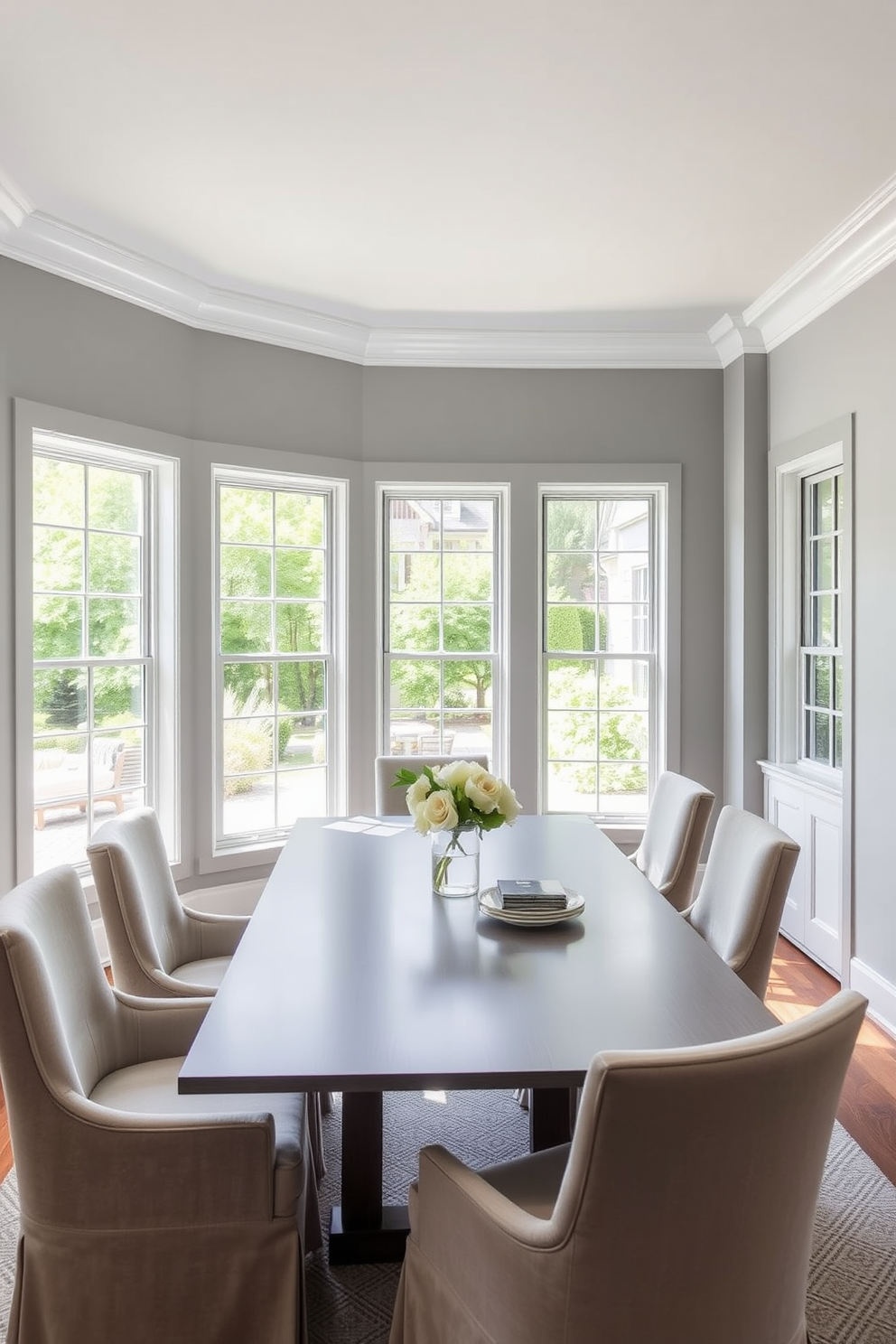 A bright and airy dining space featuring large windows that allow natural light to flood in. The walls are painted in a soft gray hue, complemented by a sleek gray dining table surrounded by elegant upholstered chairs.