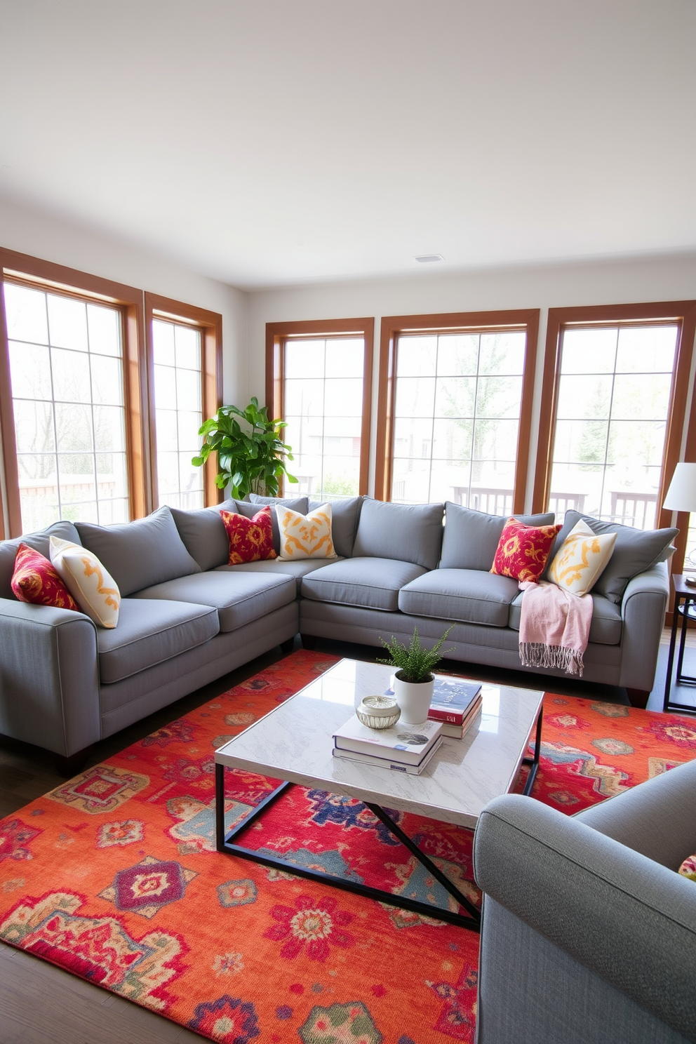 A cozy gray sectional sofa is positioned in the center of a warm family room. Colorful cushions in various patterns and textures adorn the sofa, adding a vibrant touch to the neutral palette. The walls are painted in a soft cream color, creating a bright and inviting atmosphere. A large area rug with a subtle geometric design anchors the seating area, complementing the overall decor.