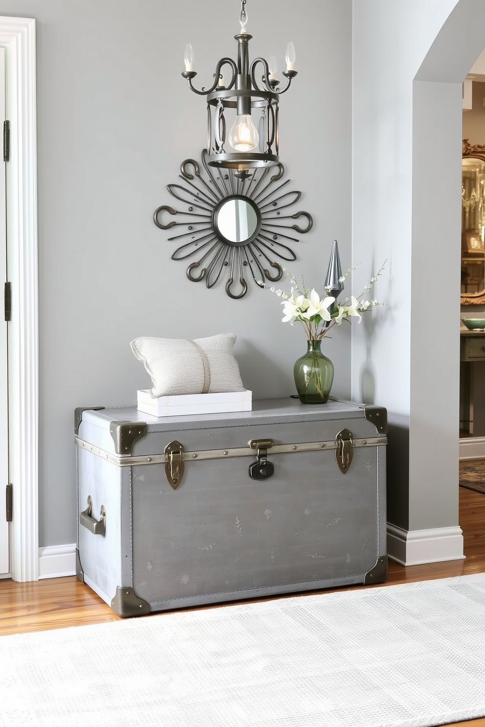 A vintage gray trunk is positioned against the wall, serving as a unique storage solution that adds character to the space. The trunk features intricate metal detailing and is complemented by a soft, neutral area rug beneath it. The foyer is designed with a blend of vintage and modern elements, showcasing a gray color palette that creates a welcoming atmosphere. A statement light fixture hangs above, illuminating the space and highlighting the elegant wall decor.