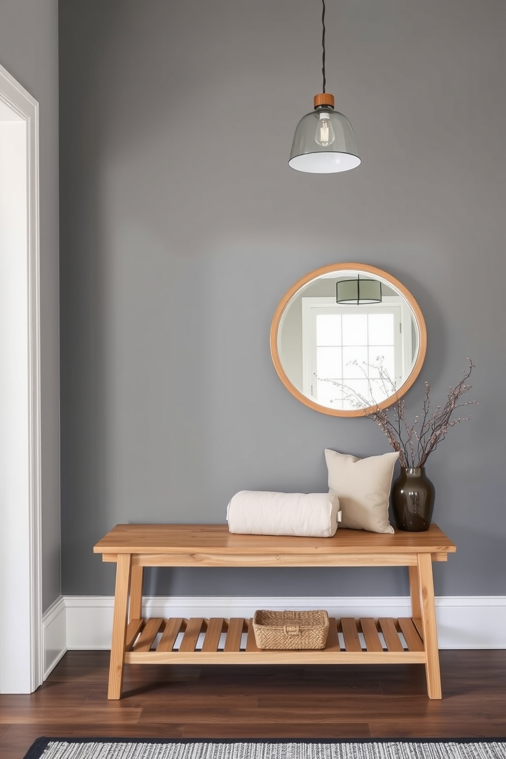 Natural wood elements to warm gray space. This foyer features a sleek gray accent wall complemented by a natural wood bench with plush cushions. A large round mirror with a wooden frame hangs above the bench, reflecting the soft light from a stylish pendant lamp. The floor is adorned with a textured rug that adds warmth and comfort to the space.