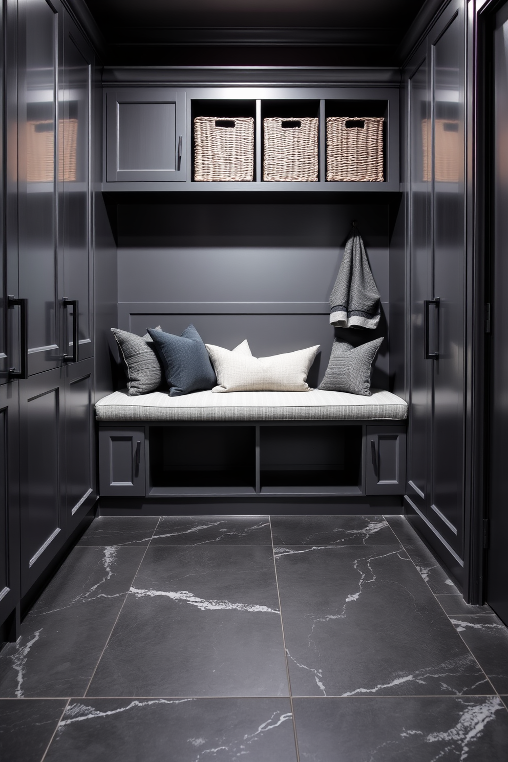 A dramatic gray and black mudroom featuring sleek cabinetry with matte black hardware. The walls are painted in a deep charcoal gray, while the floor showcases large black tiles with subtle gray veining. A stylish bench upholstered in a textured gray fabric sits against the wall, complemented by decorative pillows in varying shades of gray. Shelving above the bench holds neatly arranged baskets for storage, adding both functionality and elegance.